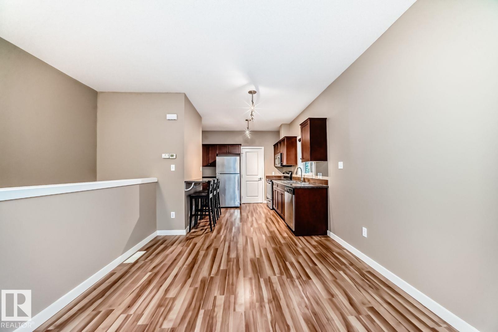 Open-concept living area featuring wood-finish flooring, a neutral color palette, and a functional kitchen with dark cabinetry, stainless steel appliances, and track lighting - 4 11917 103 Street, Edmonton, AB