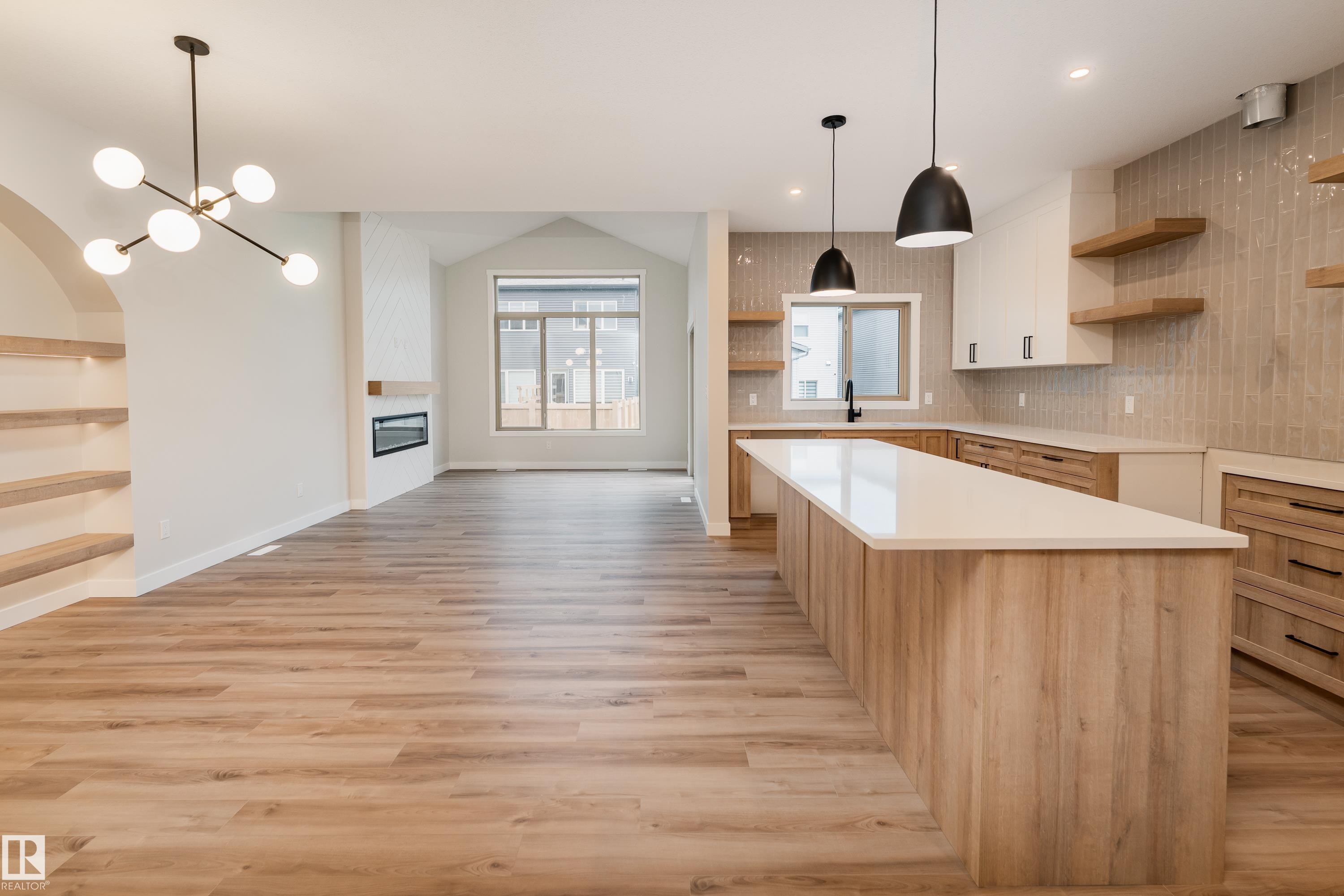 Spacious open-concept living area featuring wood-finish flooring, a contemporary kitchen island with a light-colored countertop, minimalist light fixtures, an electric fireplace with a floating mantel, and an arched built-in shelving unit - 115 Garneau Gate, Spruce Grove, AB