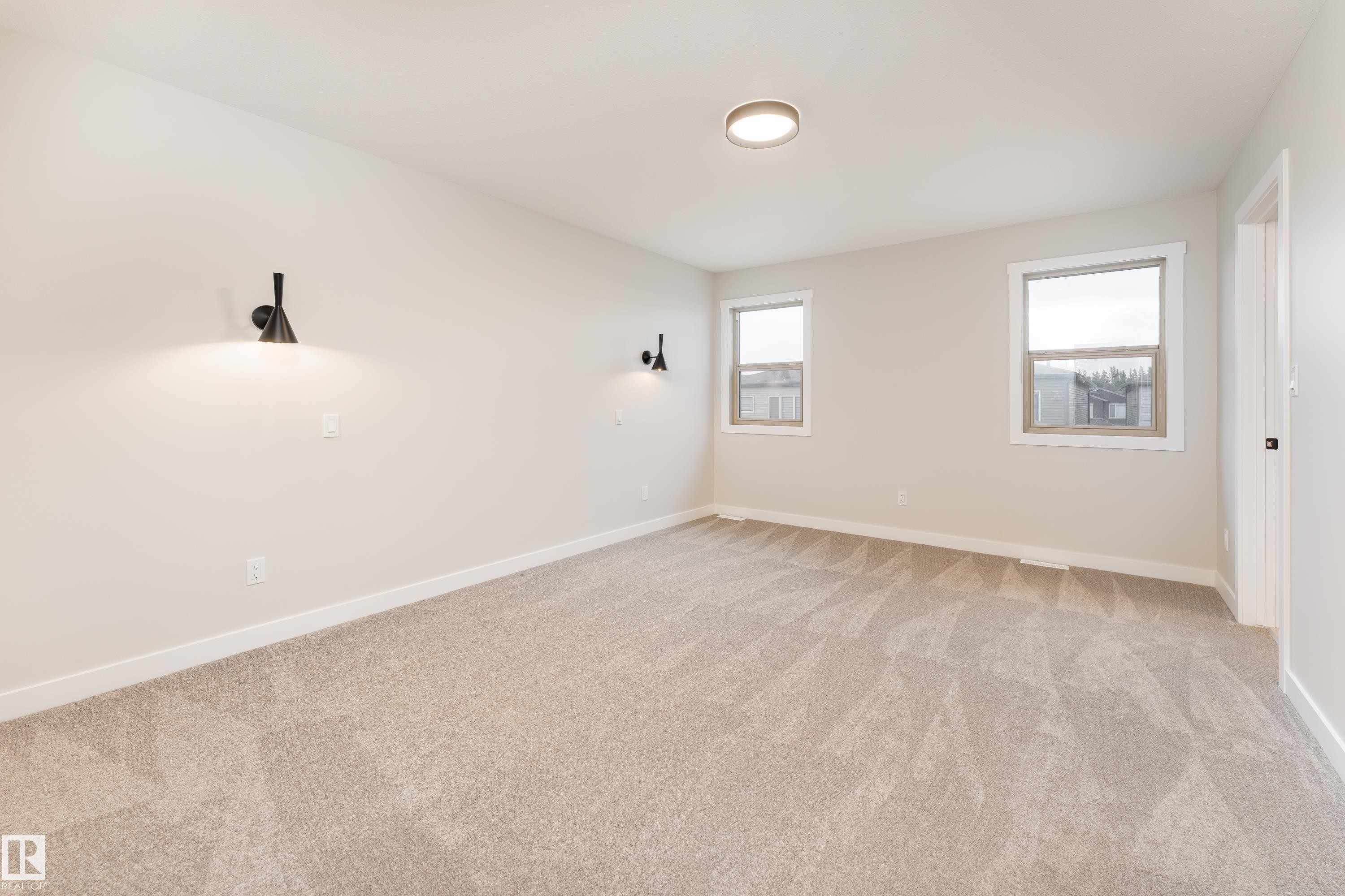 Spacious room featuring neutral carpeting, light-toned wall paint, two windows with white trim, a flush-mount ceiling light, and two wall sconces providing accent lighting - 115 Garneau Gate, Spruce Grove, AB
