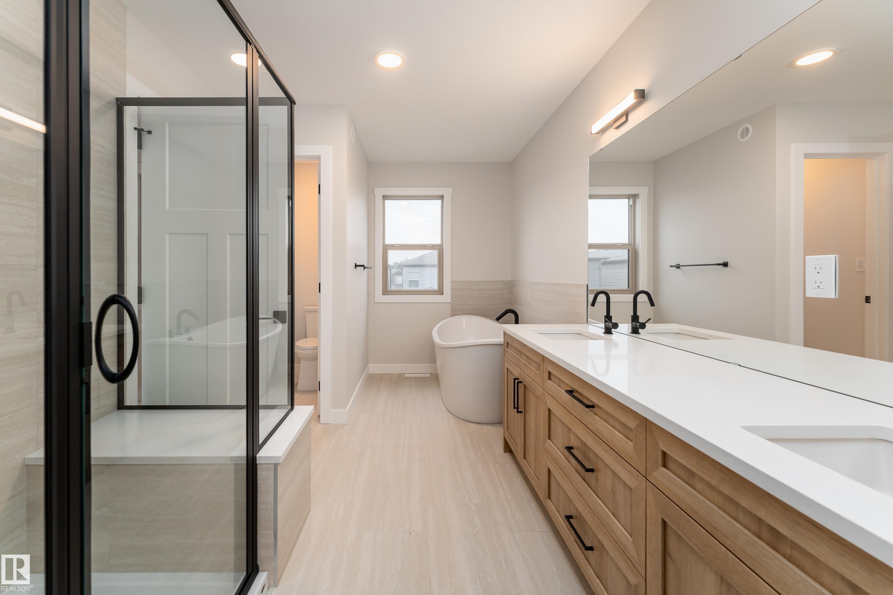Spacious bathroom featuring a dual vanity with wood-finish cabinetry and white countertops, a freestanding soaking tub, a glass-enclosed shower with a bench, and light-toned tile flooring - 115 Garneau Gate, Spruce Grove, AB