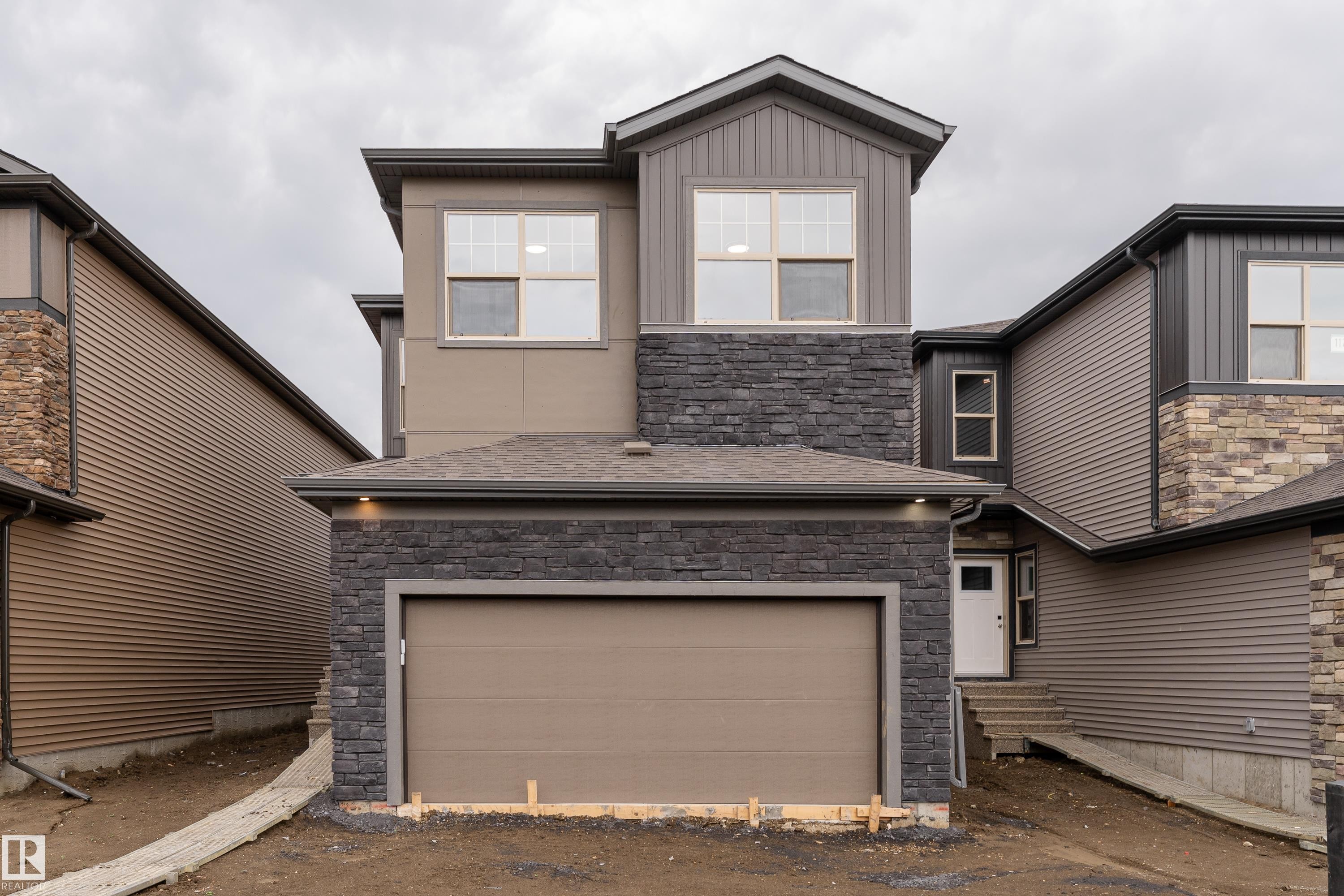 Modern exterior featuring multi-toned siding, dark stone accents, an attached garage, a contemporary entry door, and a prominent second-story window - 115 Garneau Gate, Spruce Grove, AB