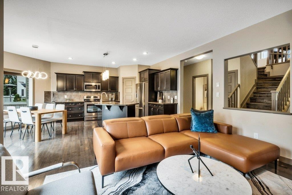 Open-concept living space featuring wood-finish flooring, a contemporary kitchen with dark cabinetry and stainless steel appliances, a dining area with modern pendant lighting, and a visible staircase with wood treads and railings - 1107 Chahley Crest, Edmonton, AB