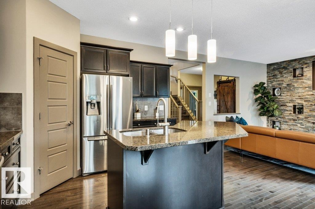 Open-concept kitchen featuring an island with a built-in sink, stainless steel appliances, dark cabinetry, and stone-finish countertops - 1107 Chahley Crest, Edmonton, AB
