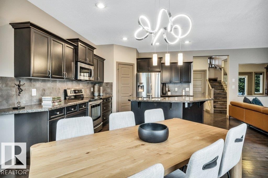 Open-concept kitchen and dining area featuring dark wood-finish cabinetry, stainless steel appliances, a tiled backsplash, and an island with pendant lighting - 1107 Chahley Crest, Edmonton, AB