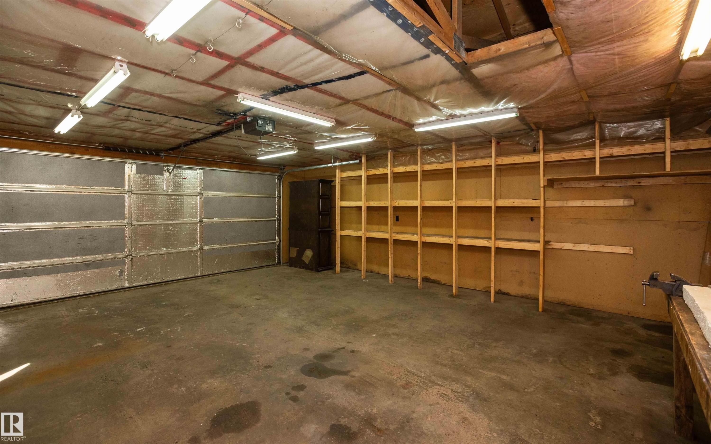 Spacious garage featuring a concrete floor, an insulated overhead door, multiple fluorescent light fixtures, exposed ceiling beams, and extensive built-in wooden shelving for storage - 4414 54 Avenue, Barrhead, AB - Indoor Photo Showing Garage