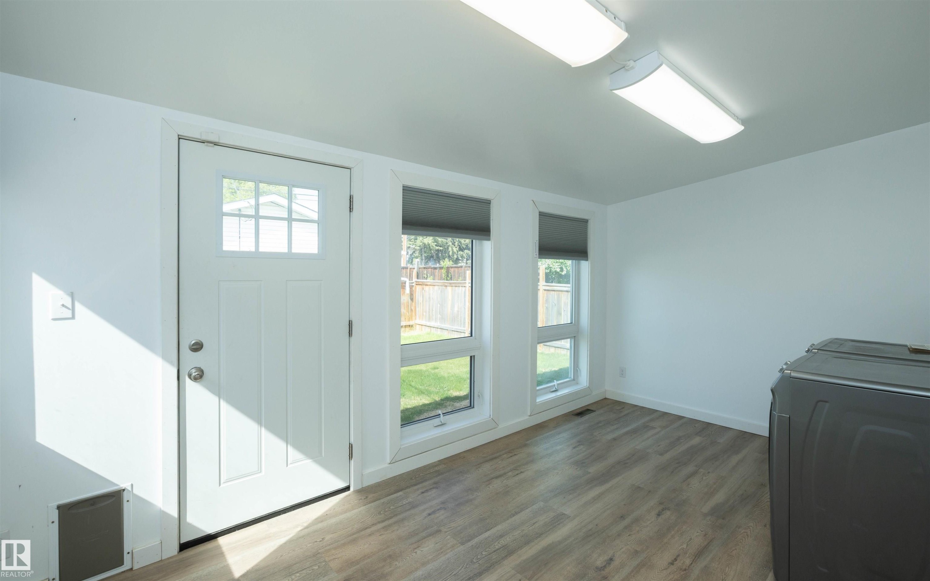 Interior space featuring wood-finish flooring, two large windows with roller shades, and a rear exit door with an integrated multi-pane window - 4414 54 Avenue, Barrhead, AB - Indoor Photo Showing Other Room