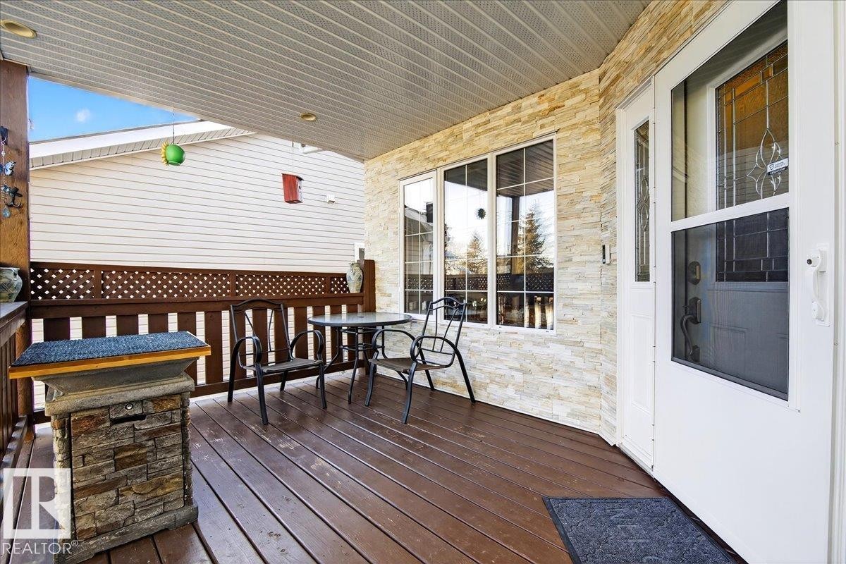 Covered porch featuring wood-finish decking, stone-faced exterior wall accents, a white storm door with decorative glass, white vinyl siding, and a lattice privacy screen - 5019 48 Avenue, Chipman, AB - Outdoor With Deck Patio Veranda With Exterior