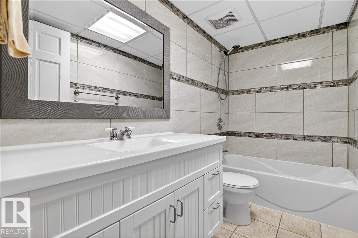 Bathroom featuring a white vanity with integrated sink, a framed rectangular mirror, a bathtub with shower, and tiled walls with decorative mosaic accents - 5019 48 Avenue, Chipman, AB - Indoor Photo Showing Bathroom
