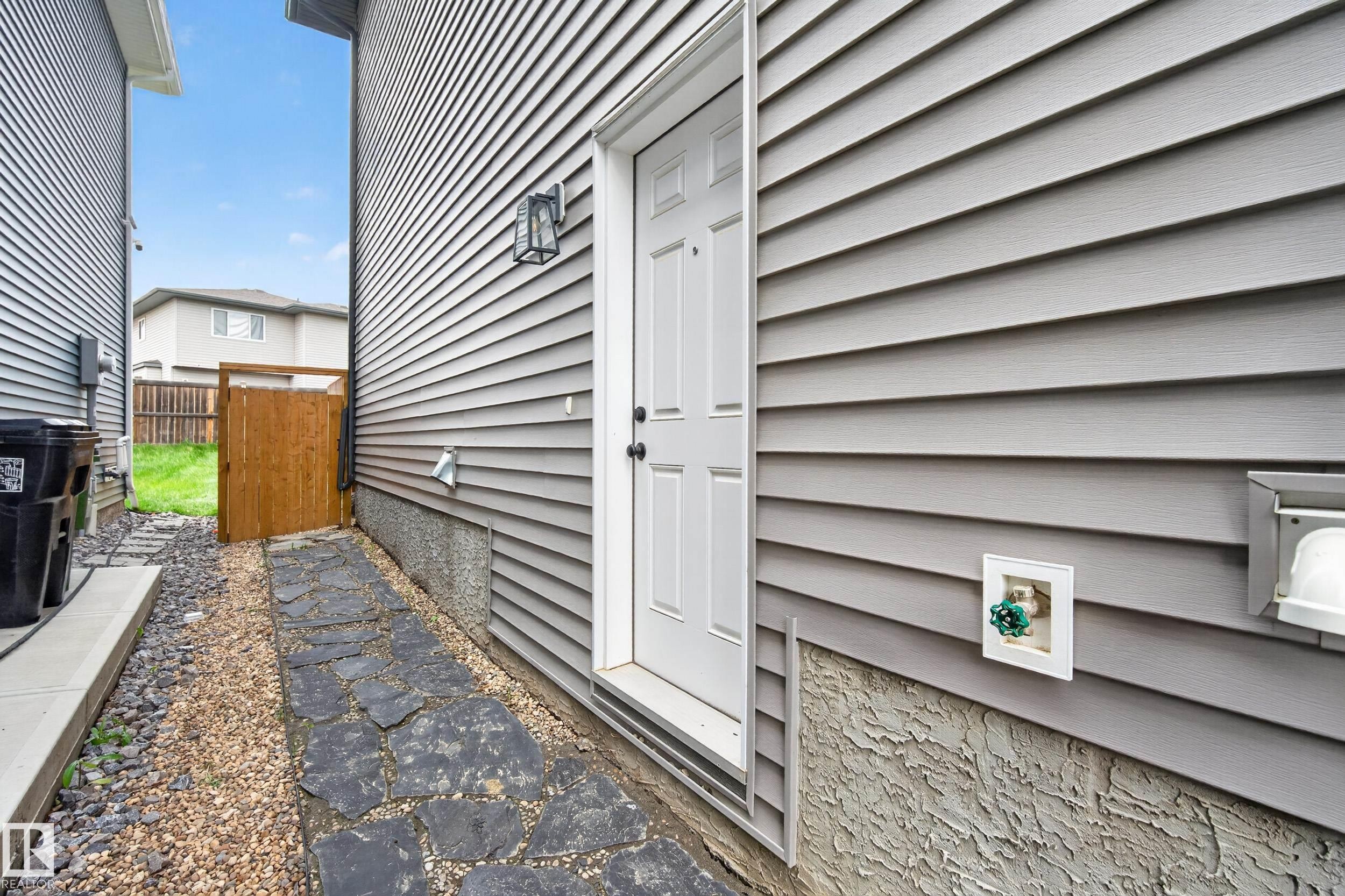 Grey horizontal siding exterior featuring a white paneled door with black hardware, a black wall-mounted lantern light fixture, and a stone slab pathway - 22836 94 Avenue, Edmonton, AB