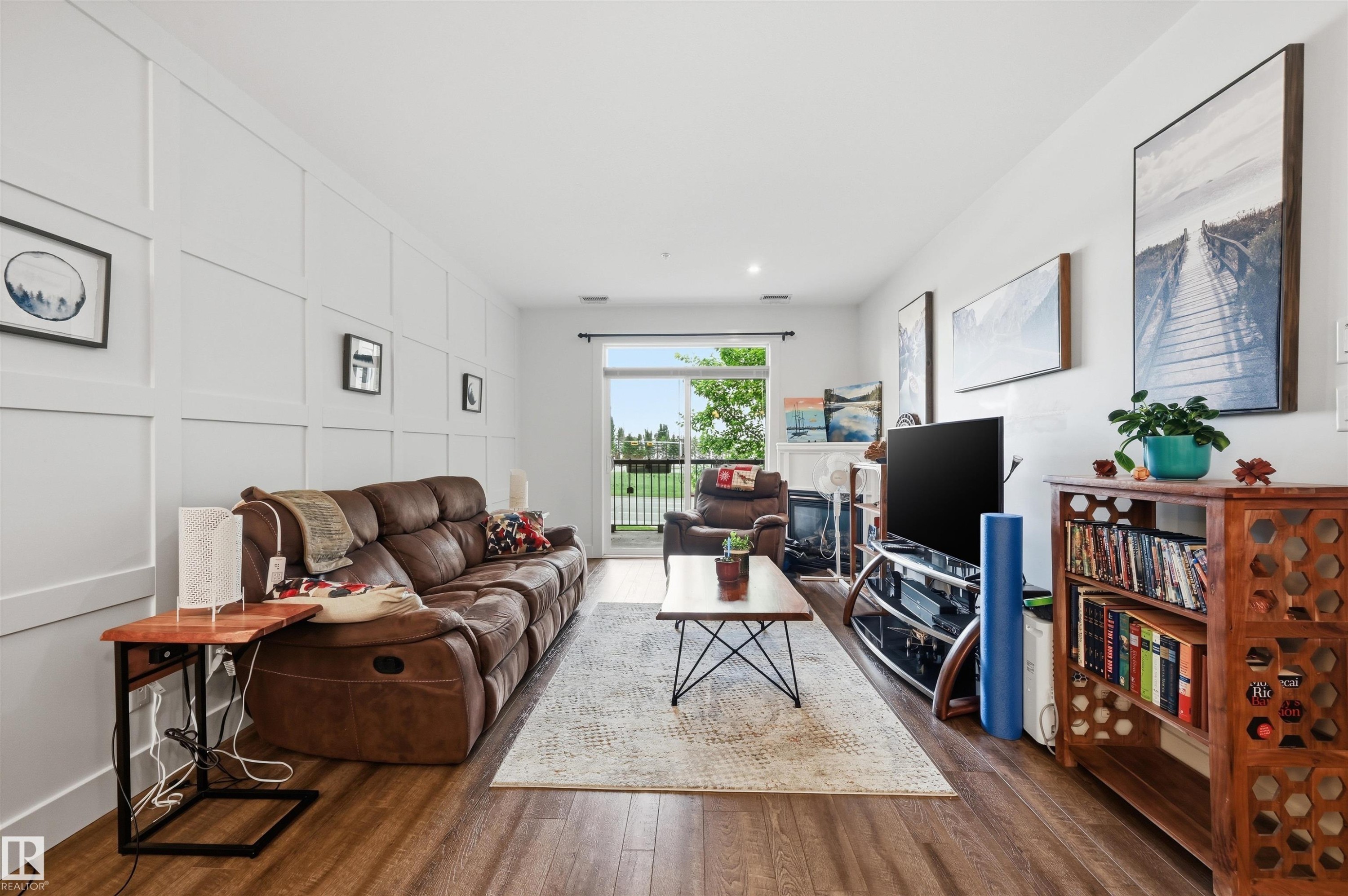 Spacious living area featuring a board and batten accent wall, wood-finish flooring, a glass sliding door, and recessed lighting - 217 2503 Hanna Crescent, Edmonton, AB