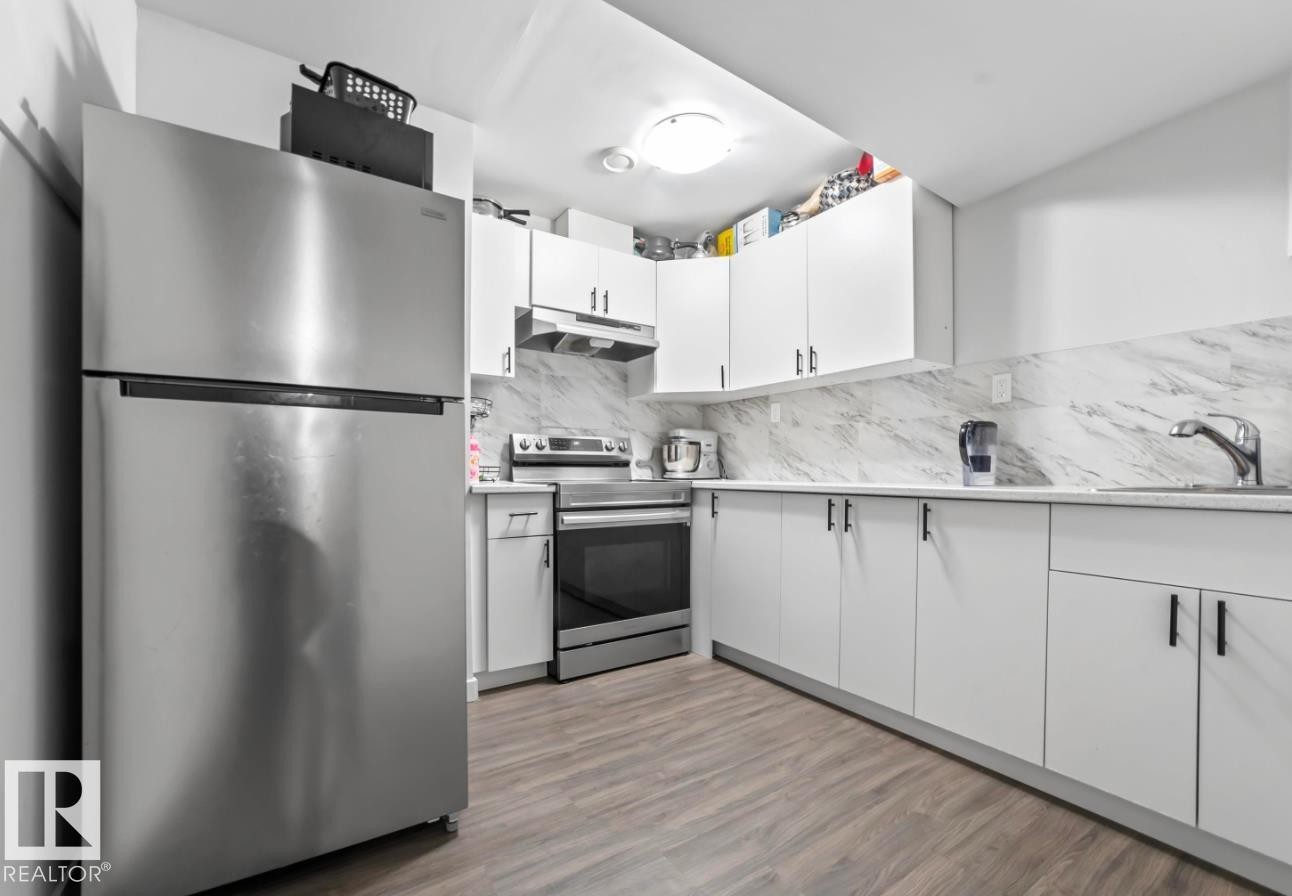 Modern kitchen featuring stainless steel appliances, white cabinetry with black hardware, stone-look countertops, marble-patterned backsplash, and wood-finish flooring - 2153 Maple Road, Edmonton, AB