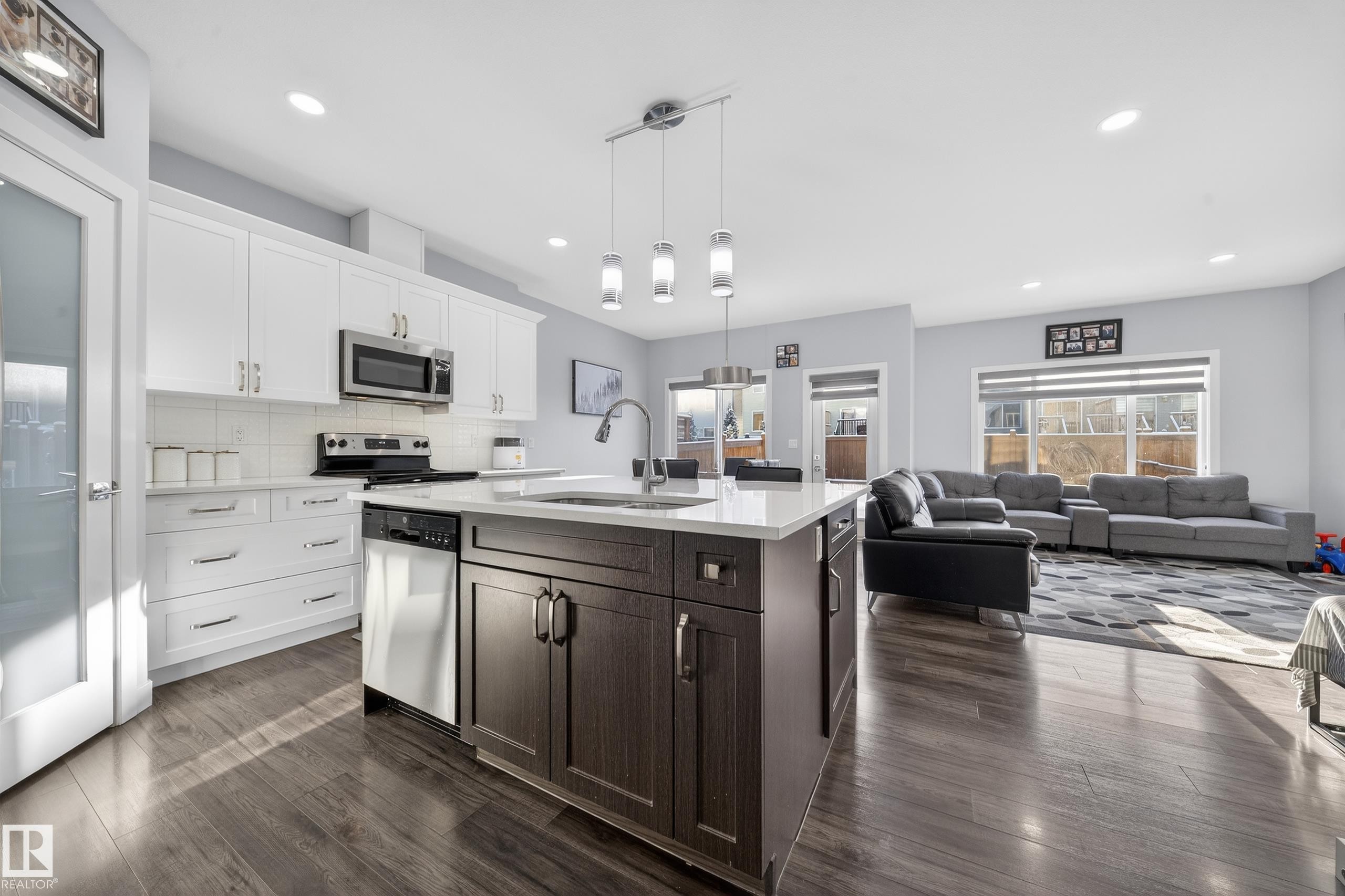 Open-concept living space featuring wood-finish flooring, a kitchen island with pendant lighting, white cabinetry, stainless steel appliances, and large windows - 2084 Graydon Hill, Edmonton, AB