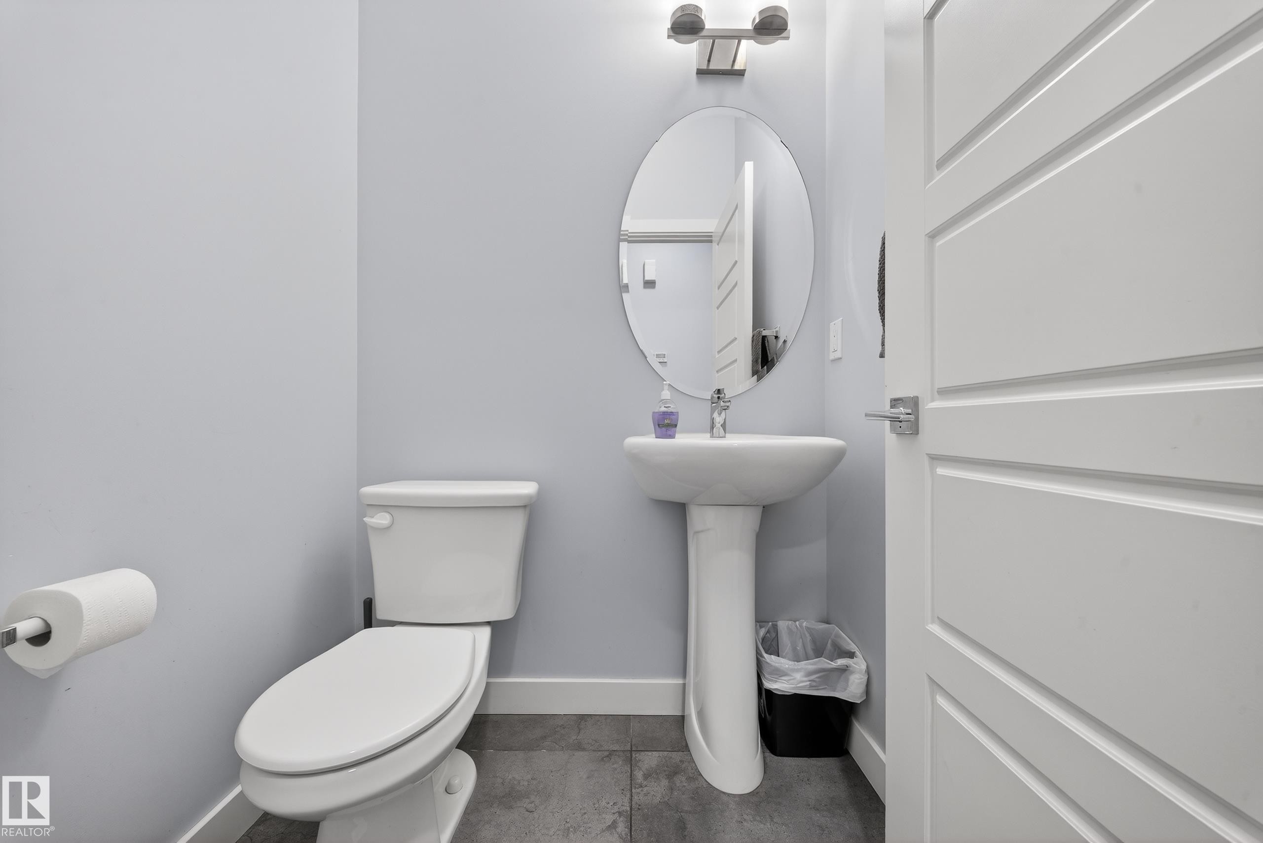 Modern powder room featuring a pedestal sink, oval mirror, wall-mounted light fixture, and contemporary paneled door - 2084 Graydon Hill, Edmonton, AB