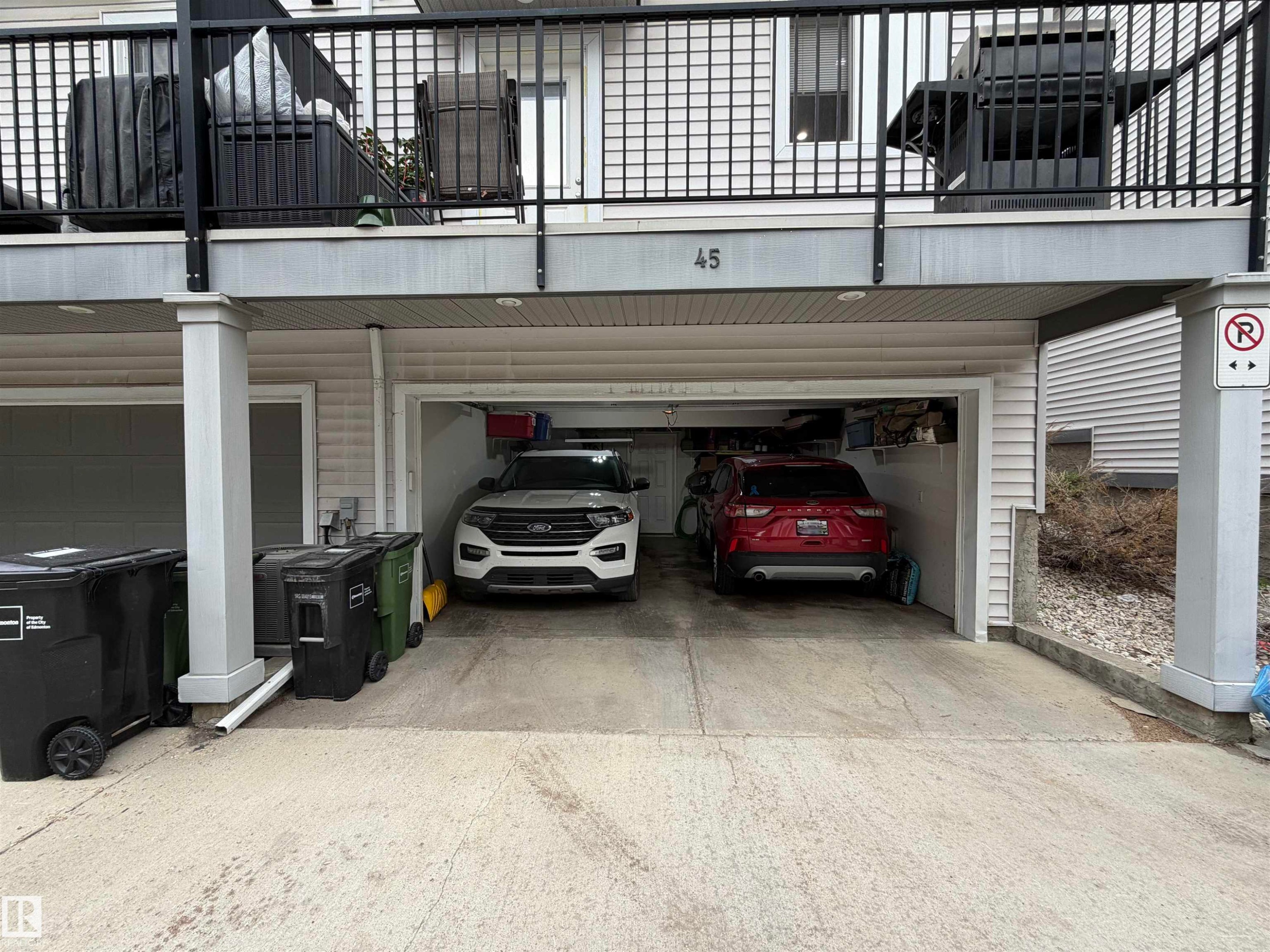Two-car garage with overhead lighting, featuring a concrete driveway and an upper-level balcony with black railings - 45 6075 Schonsee Way, Edmonton, AB