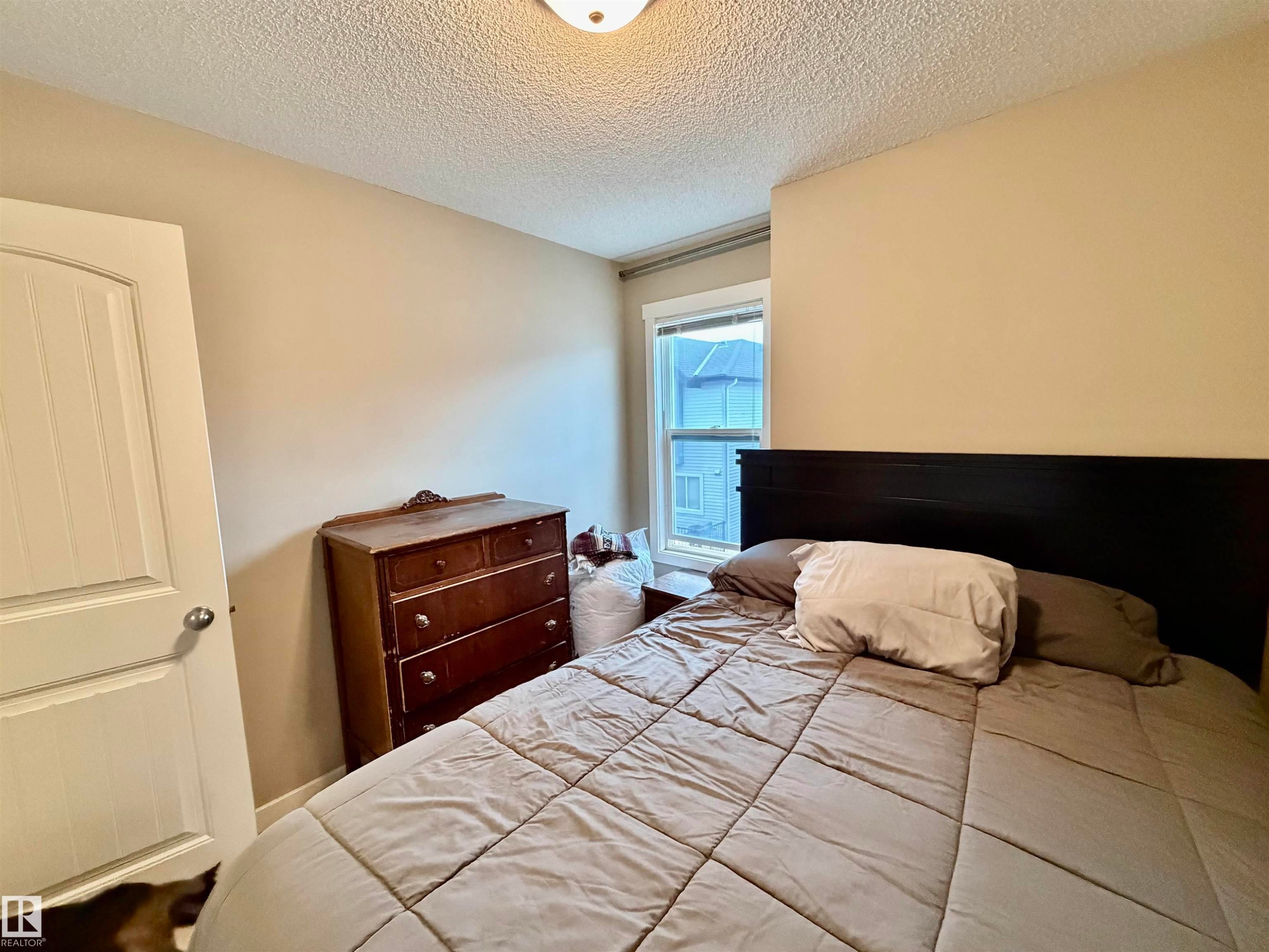Neutral-toned bedroom featuring a single window with white trim, a wood-finish door, and textured ceiling - 45 6075 Schonsee Way, Edmonton, AB