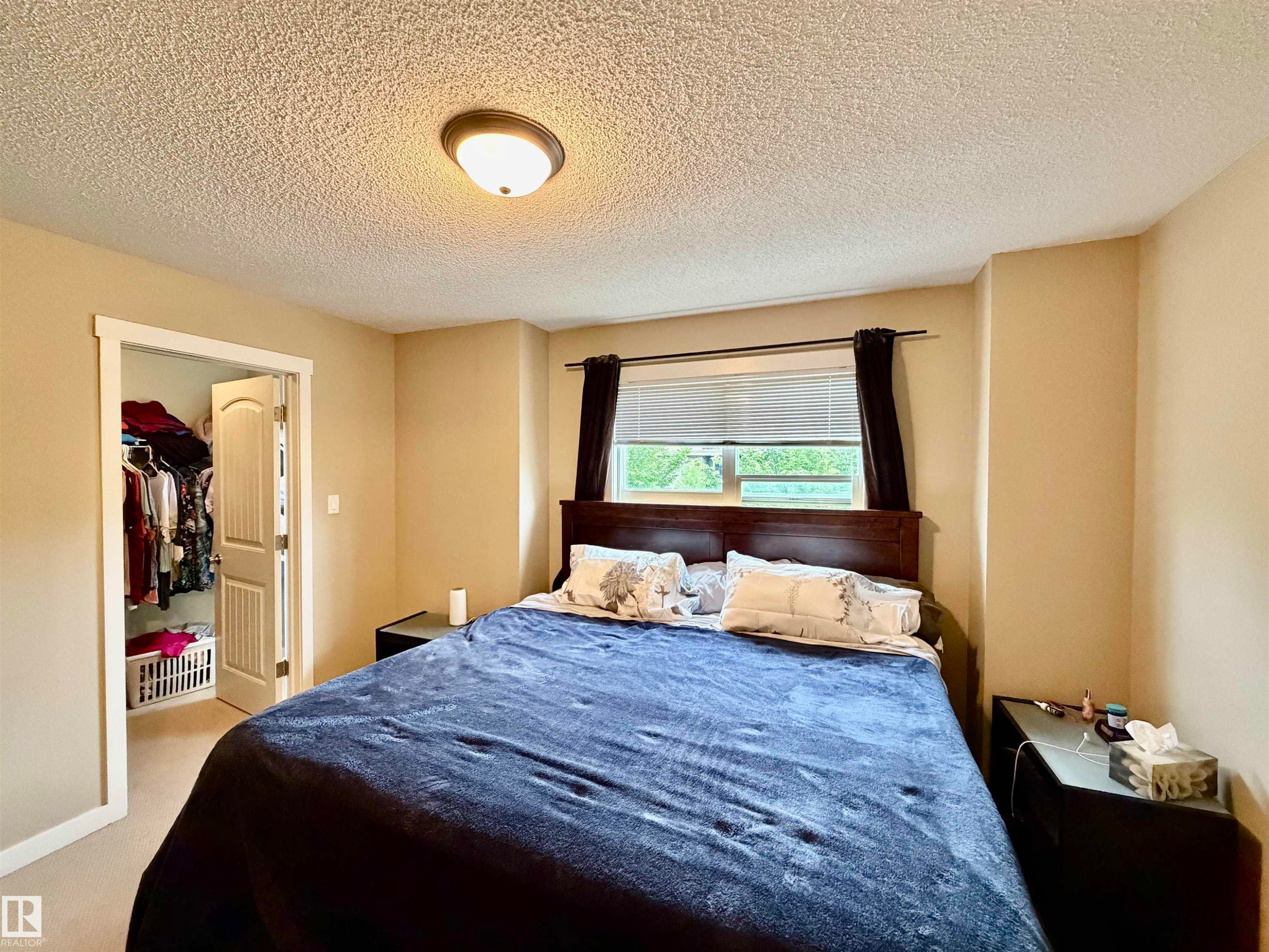 Bedroom featuring a window with blinds, wood-finish headboard, flush-mount ceiling light fixture, and an open closet with bi-fold doors - 45 6075 Schonsee Way, Edmonton, AB