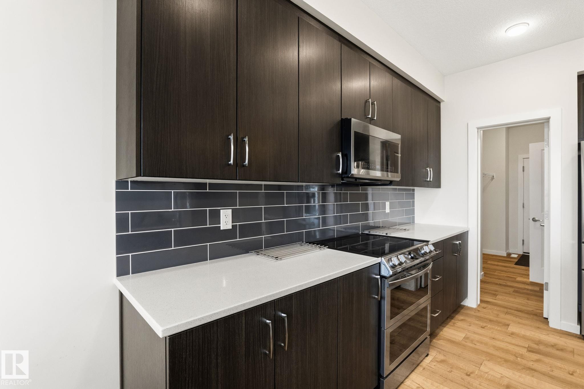 Modern kitchen featuring dark wood-finish cabinetry, light-toned countertops, a full subway tile backsplash, stainless steel appliances, and wood-look flooring - 20011 26, Edmonton, AB