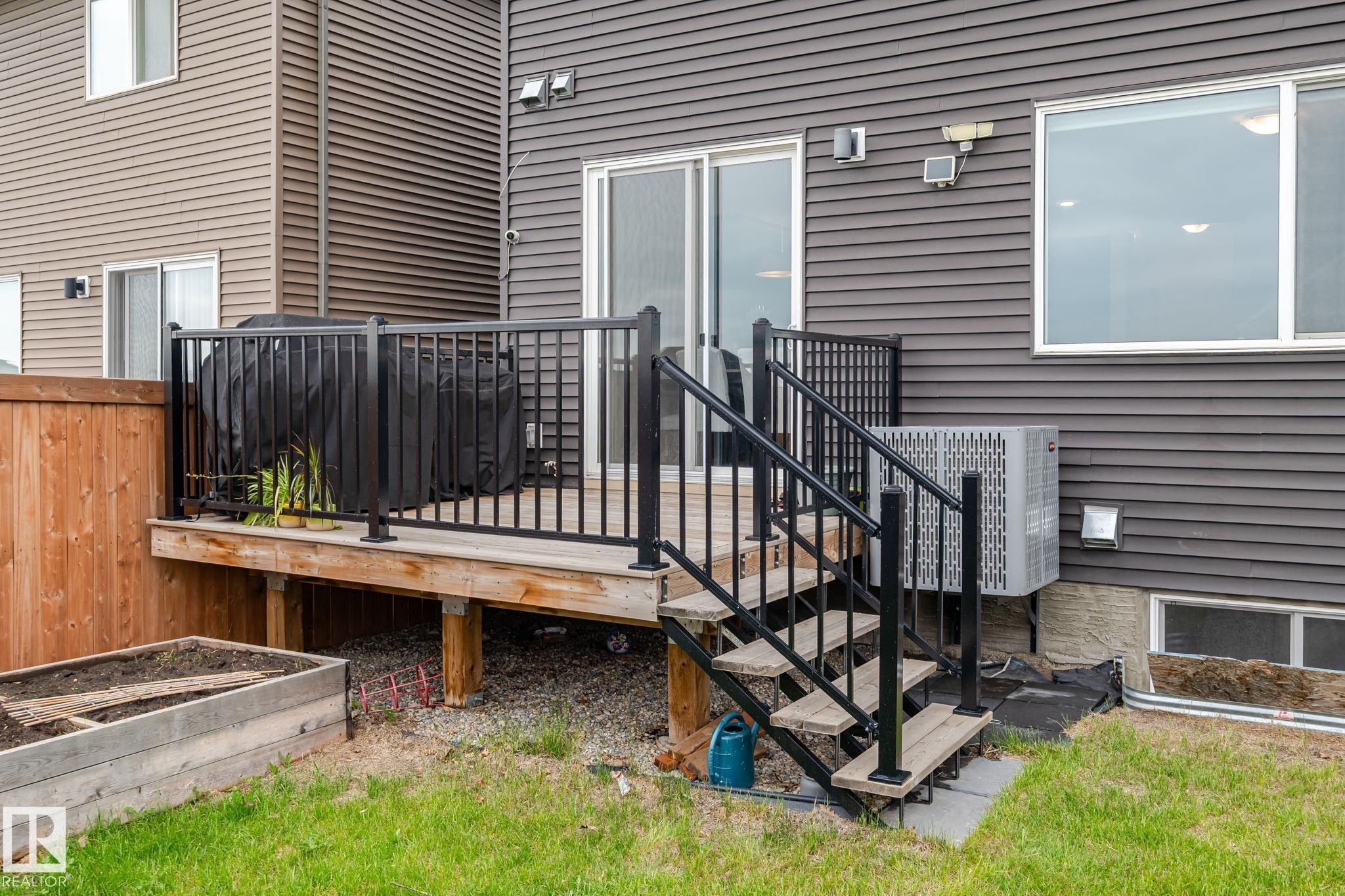 Rear exterior featuring a wooden deck with black metal railings, a sliding glass door, a large window, a raised garden bed, and a grass lawn - 20011 26, Edmonton, AB