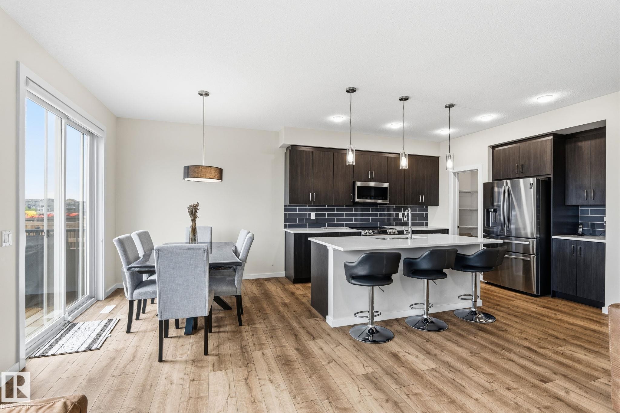 Open-concept kitchen and dining area featuring wood-finish flooring, dark wood cabinetry, stainless steel appliances, a kitchen island with seating, and sliding glass doors - 20011 26, Edmonton, AB