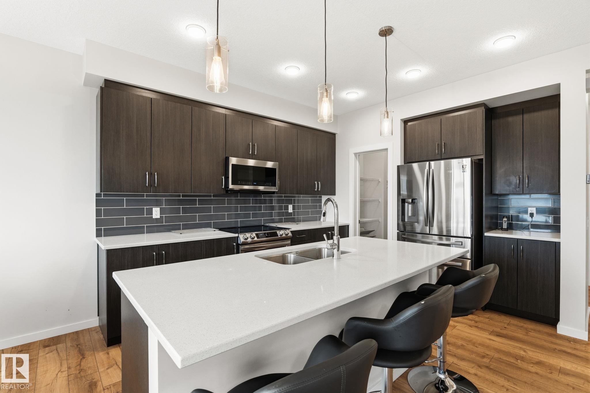 Kitchen featuring a central island with white countertop, dark wood-finish cabinetry, stainless steel appliances, and a gray subway tile backsplash - 20011 26, Edmonton, AB