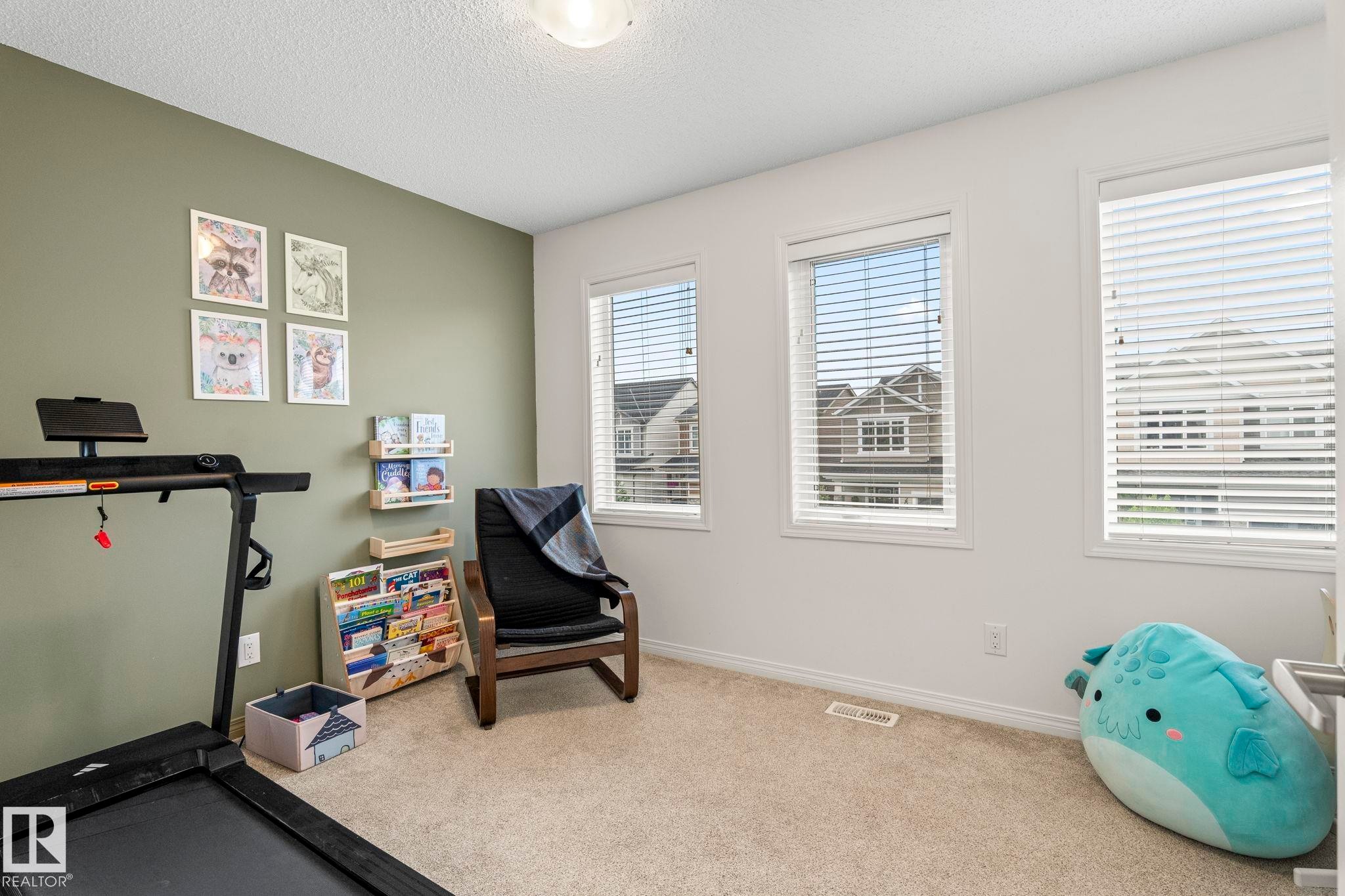 Room featuring light beige carpet flooring, multiple windows with blinds, a prominent accent wall in a muted green tone, and a flush-mount ceiling light fixture - 20227 17Ave, Edmonton, AB