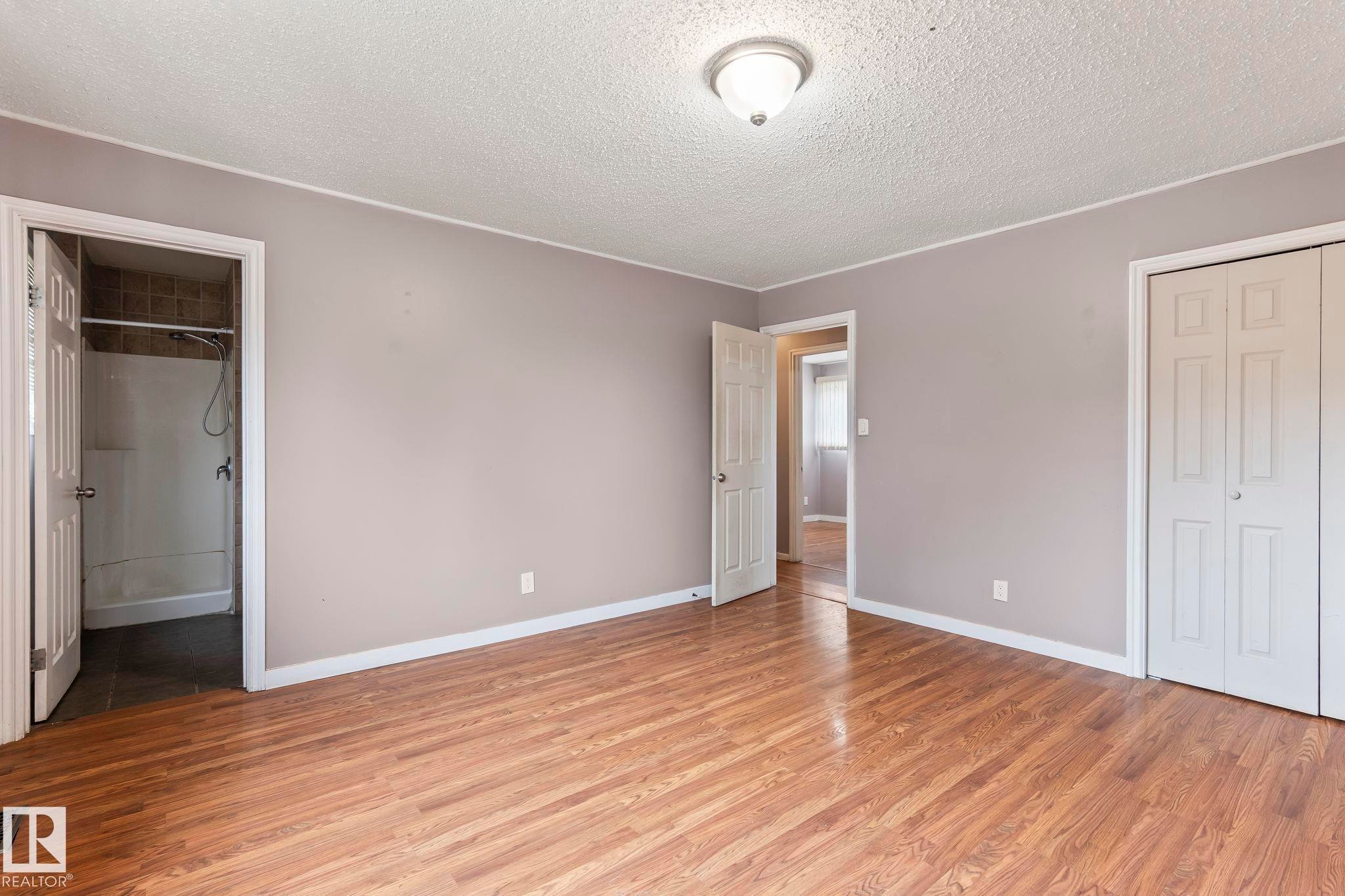 Wood-finish flooring throughout, featuring a neutral color palette, ceiling-mounted light fixture, bi-fold closet doors, and an en-suite bathroom with a shower - 2045 46St, Edmonton, AB