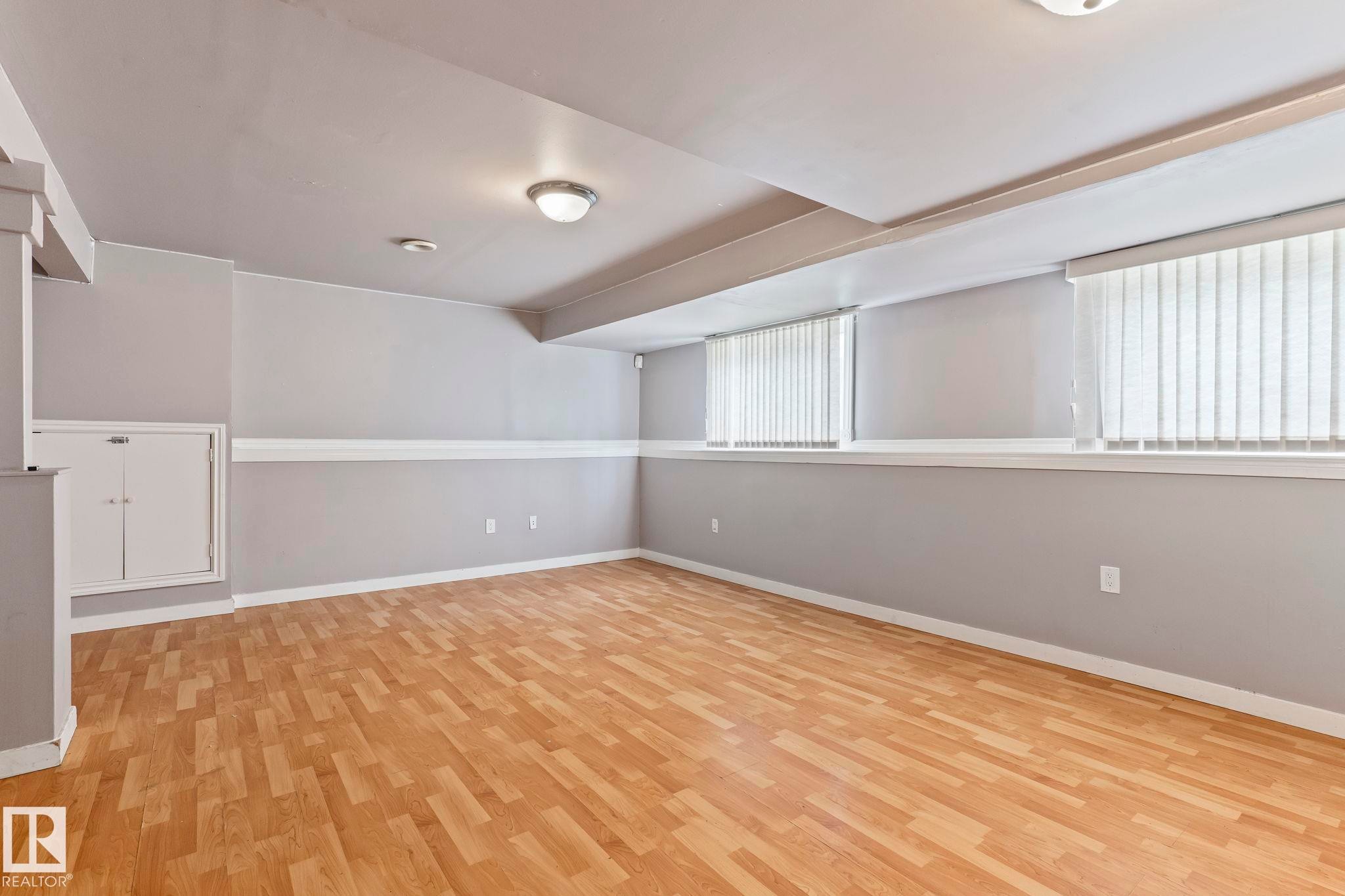 Spacious room featuring wood-finish flooring, light gray walls with a white chair rail, two windows with vertical blinds, and ceiling-mounted lighting - 2045 46St, Edmonton, AB