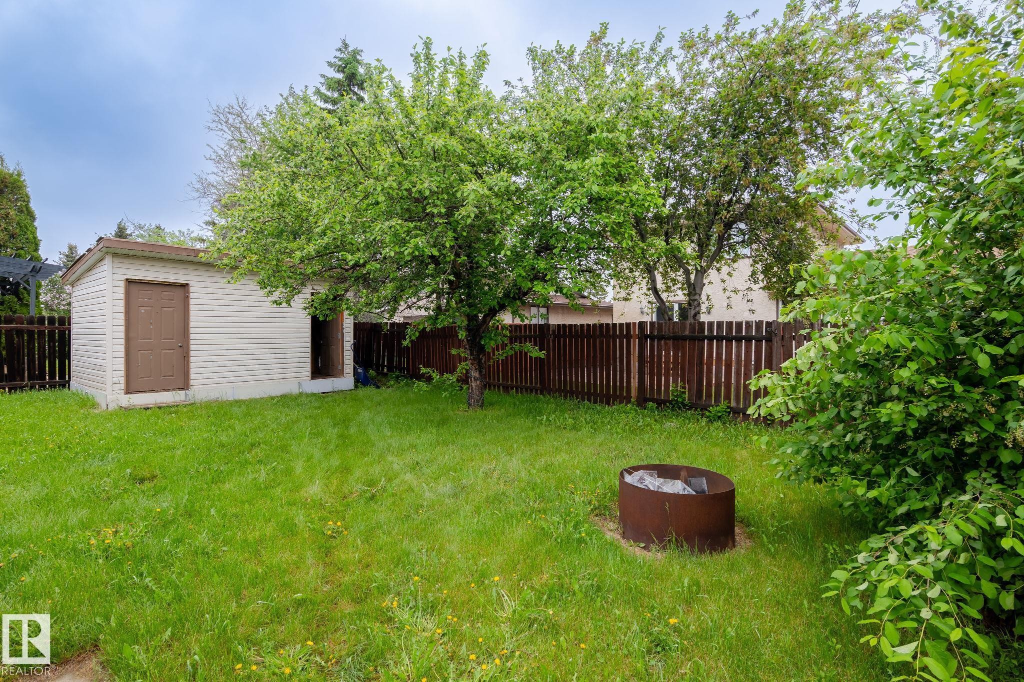 Expansive backyard featuring a detached storage shed with vinyl siding, mature shade trees, a wood privacy fence, and a built-in fire pit - 2045 46St, Edmonton, AB