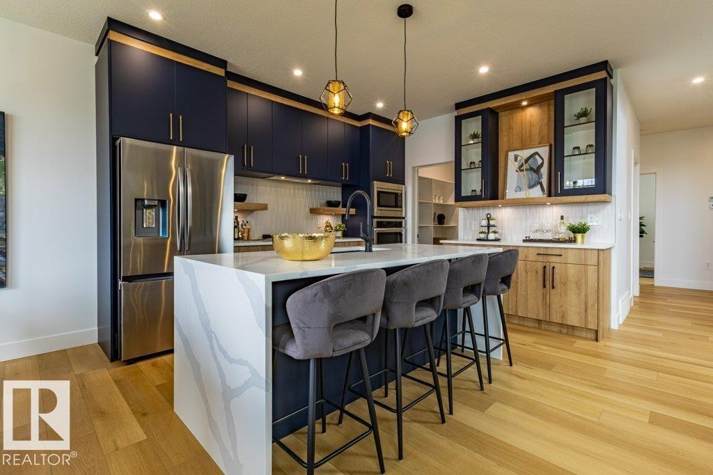 Kitchen featuring a waterfall island with stone countertop, stainless steel appliances, dark blue cabinetry, wood-finish flooring, and built-in glass display cabinets - 82 Wynn Rd, Fort Saskatchewan, AB