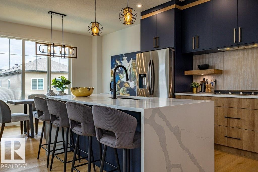Contemporary kitchen featuring a waterfall island with a light-toned countertop, contrasting dark blue cabinetry, wood-finish base cabinets, light wood-finish flooring, and stainless steel appliances - 82 Wynn Rd, Fort Saskatchewan, AB