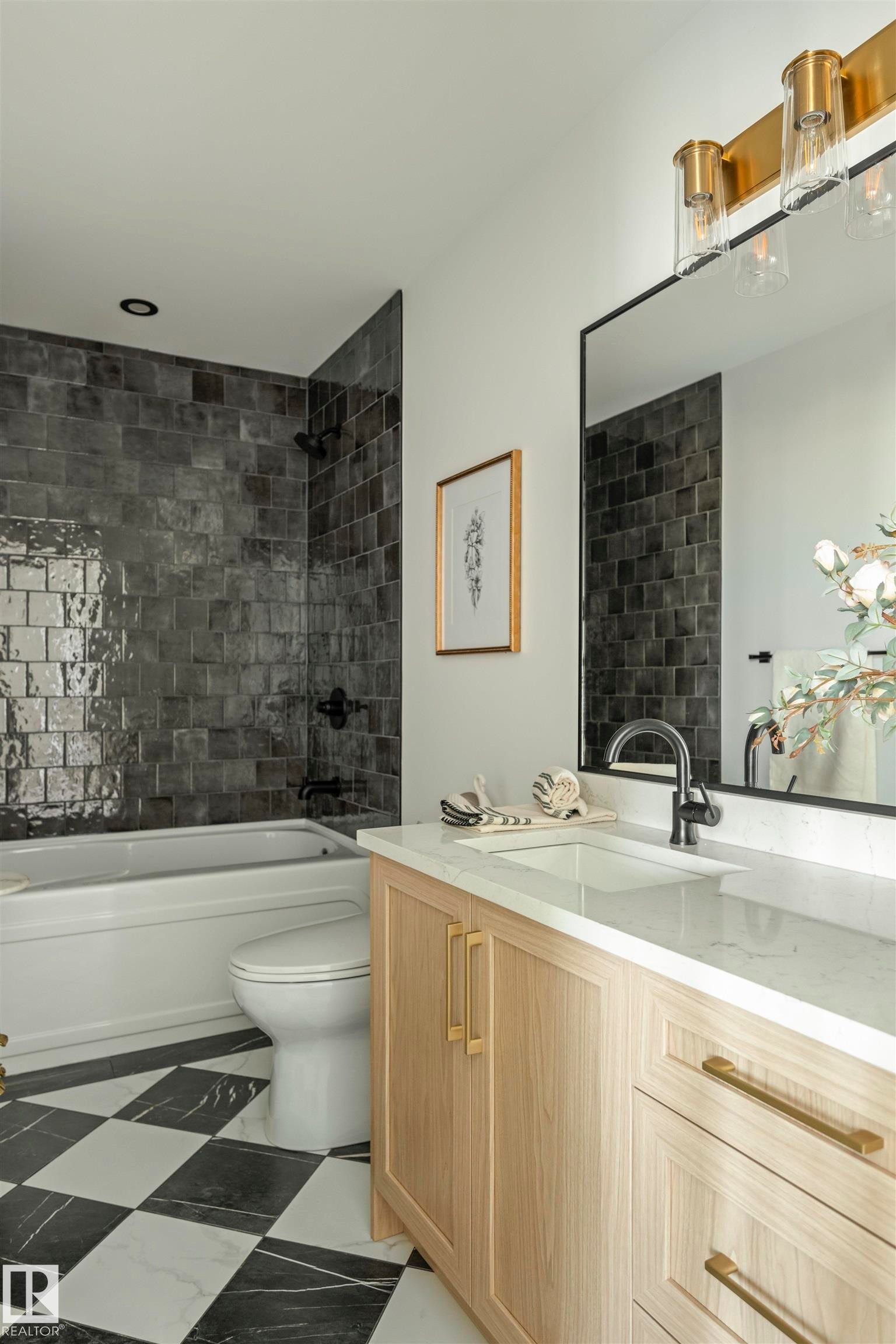 Bathroom featuring a light wood-finish vanity with a white countertop and rectangular sink, black and white checkered floor tiles, a built-in tub with dark subway tile surround, and a rectangular mirror with an overhead brass light fixture - 3329 Chickadee Drive, Edmonton, AB
