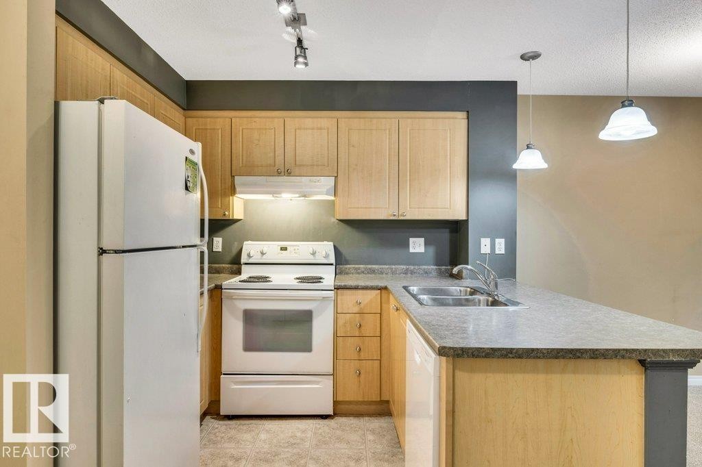 Kitchen featuring light wood cabinetry, white appliances, a double basin sink, track lighting, and pendant lighting - 116 630 Mcallister Loop, Edmonton, AB