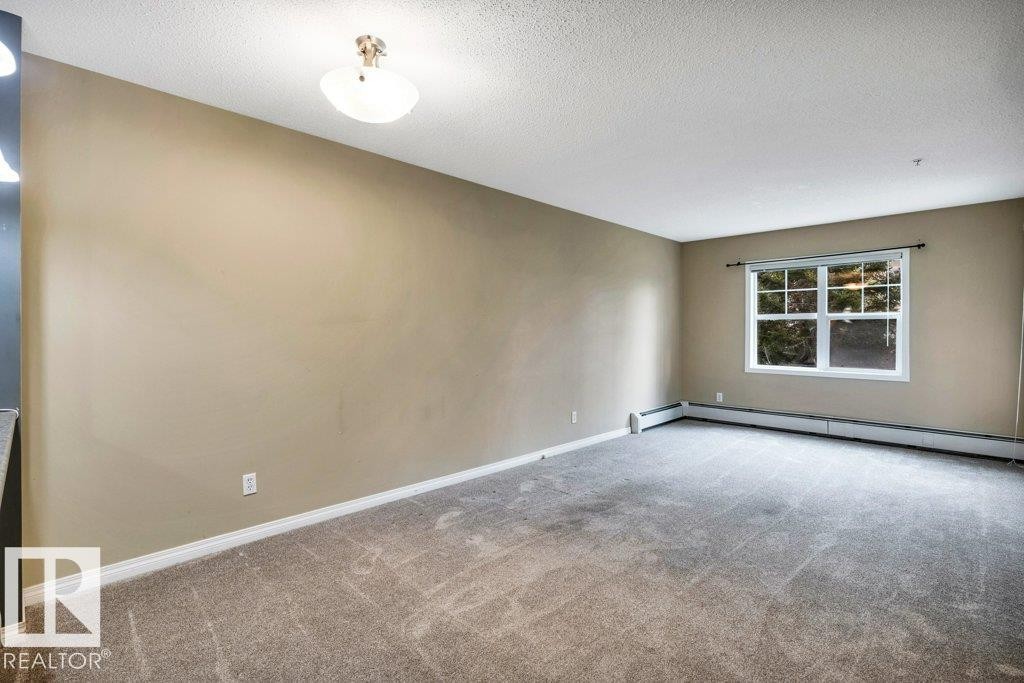 Spacious room featuring light tan walls, textured ceiling, and gray patterned carpet - 116 630 Mcallister Loop, Edmonton, AB