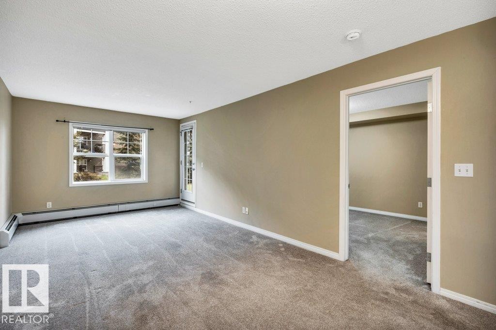 Spacious room featuring neutral-toned walls, gray carpeting, a large window, a glass-paneled door, and white trim - 116 630 Mcallister Loop, Edmonton, AB