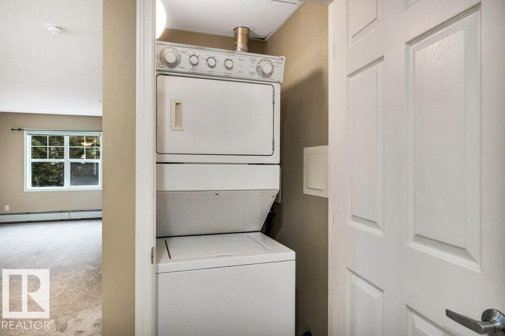 White paneled door opening to a stacked white washer and dryer unit, featuring a light tan wall and off-white carpet flooring - 116 630 Mcallister Loop, Edmonton, AB