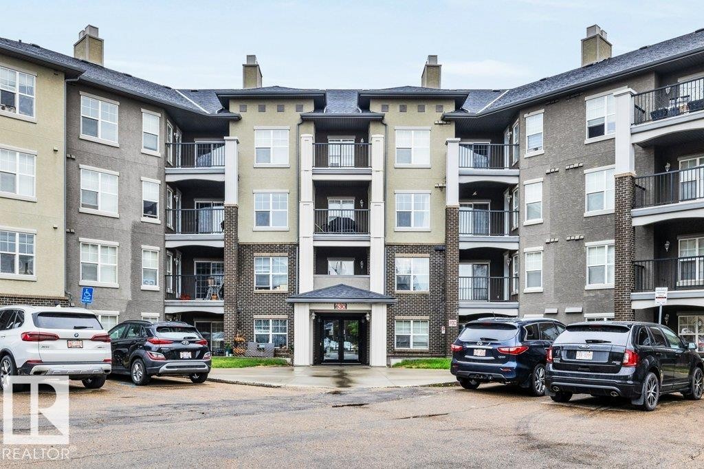Modern multi-story residential building featuring a central entrance canopy, multiple private balconies with dark railings, and a facade combining brick-finish and stucco elements - 116 630 Mcallister Loop, Edmonton, AB
