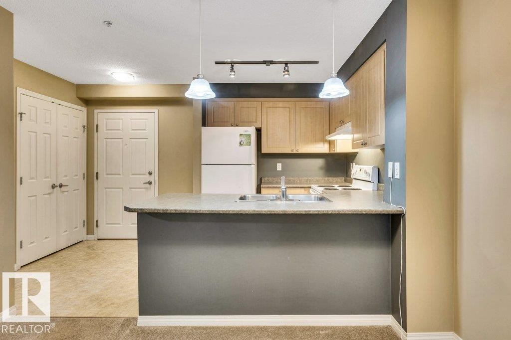 Kitchen featuring a peninsula counter with dual pendant lighting, light wood cabinetry, white appliances, and a double basin sink - 116 630 Mcallister Loop, Edmonton, AB