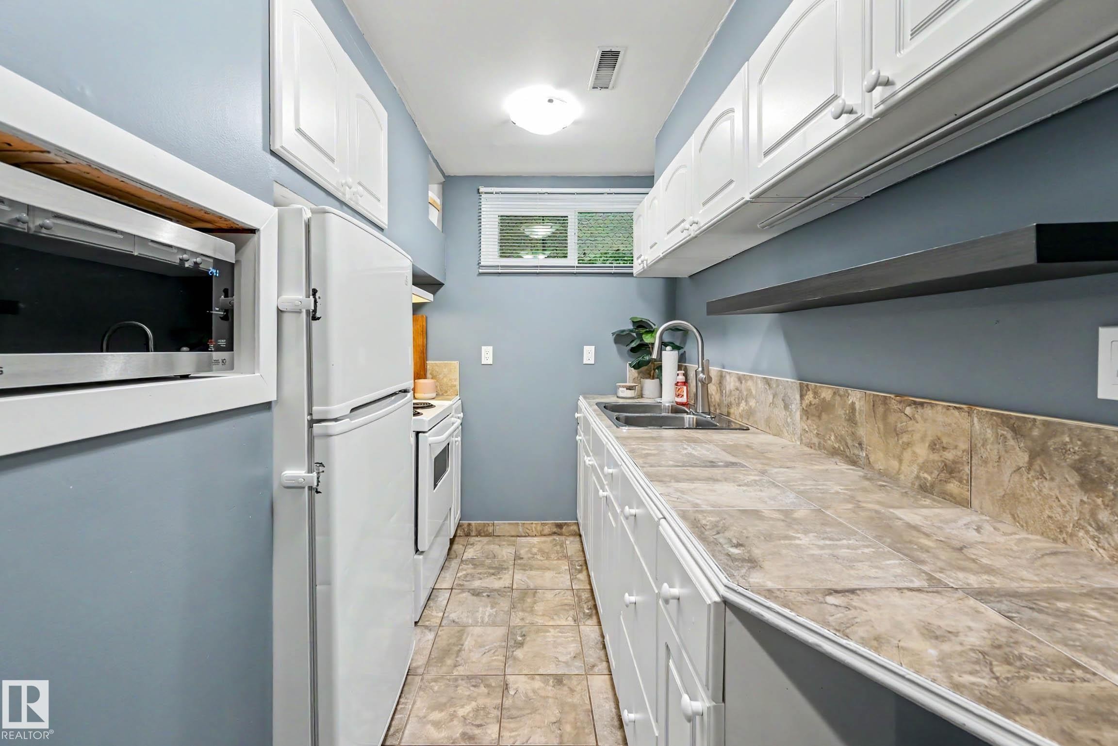 Galley kitchen featuring white cabinetry, a stainless steel sink, tiled countertops, and a tiled backsplash - 12115 40 Avenue Nw, Edmonton, AB