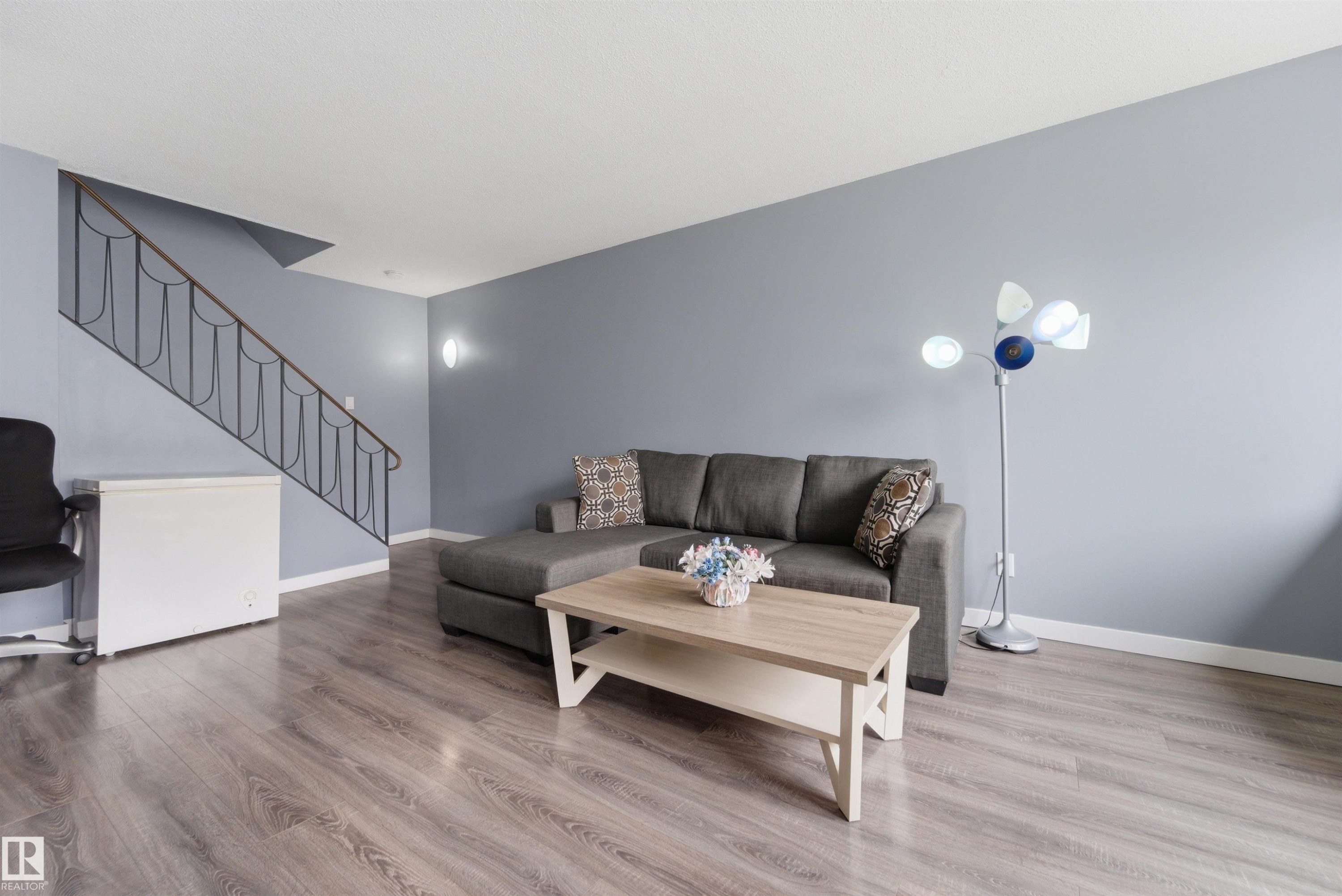 Spacious room featuring wood-finish flooring, a contemporary staircase with wrought iron railing, and neutral wall tones - #110 7805 159 St Nw, Edmonton, AB
