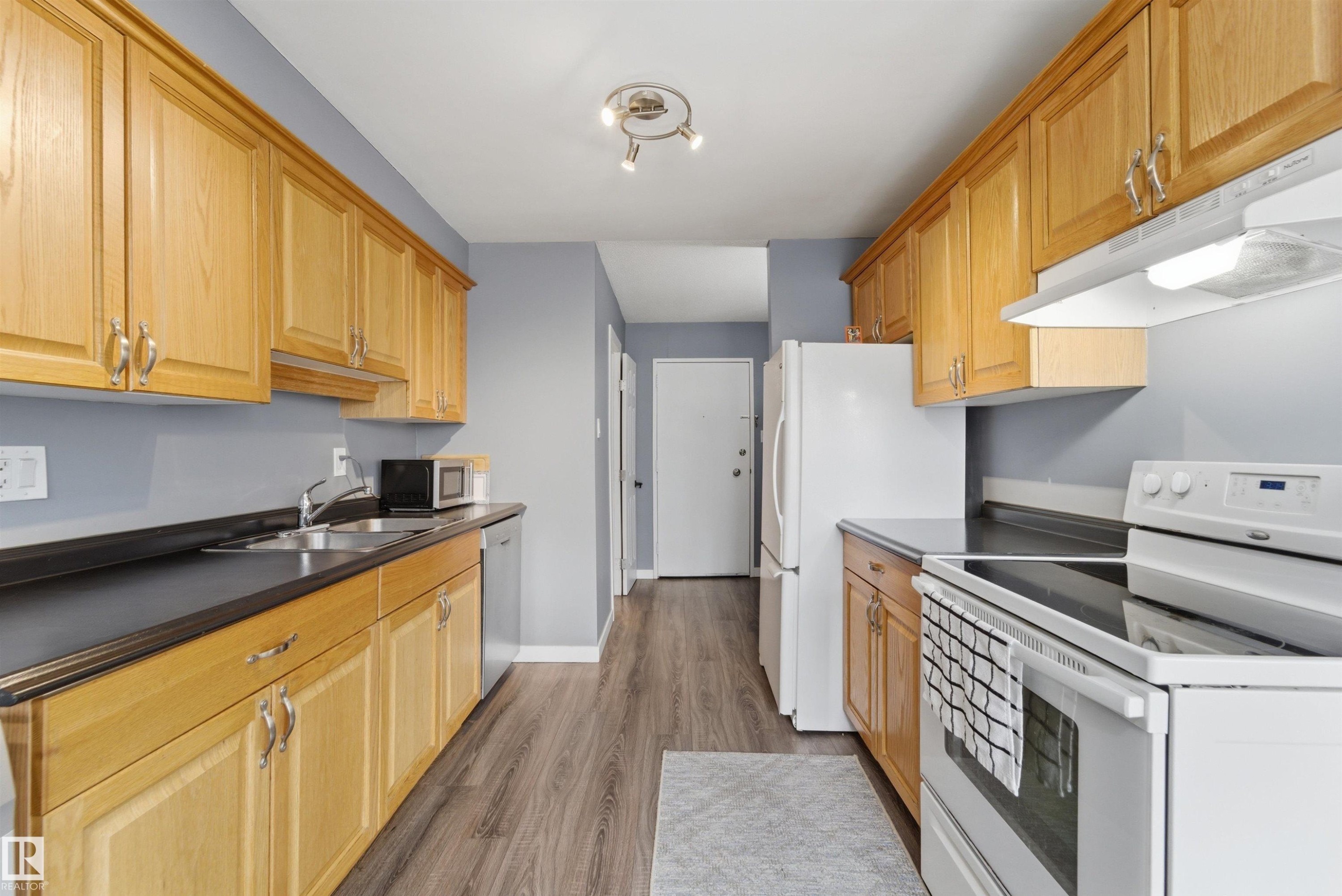 Galley kitchen featuring wood cabinetry, dark countertops, and wood-finish flooring - #110 7805 159 St Nw, Edmonton, AB