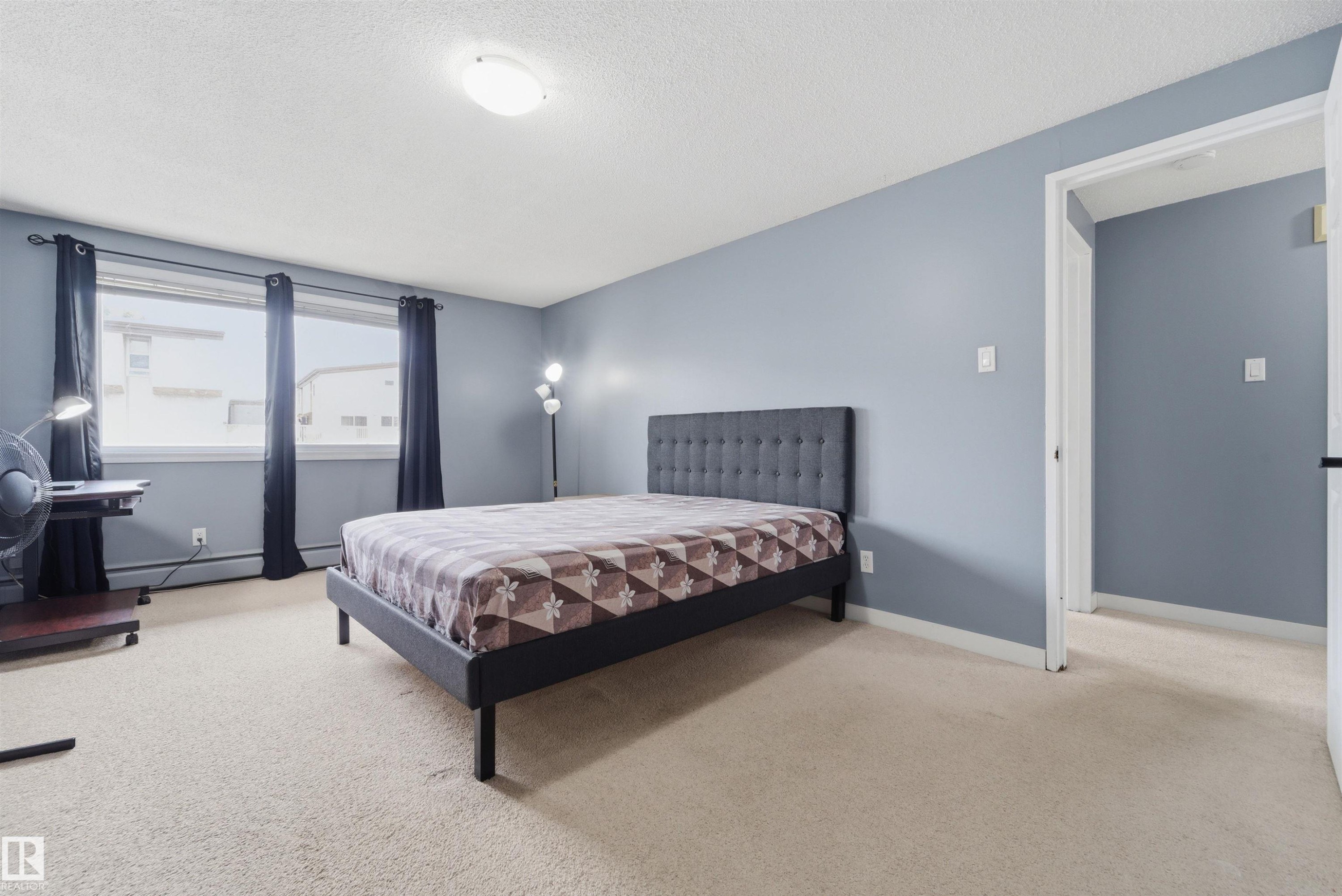 Spacious room featuring light blue walls, soft neutral carpeting, and a large window with dark drapery - #110 7805 159 St Nw, Edmonton, AB