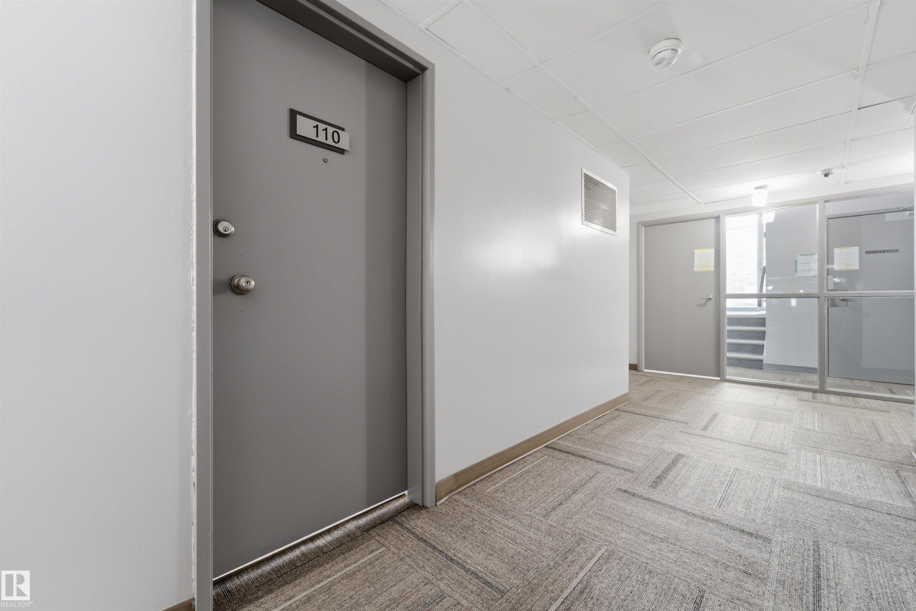 Common area hallway featuring patterned carpet flooring, white walls, and a dropped ceiling with integrated lighting - #110 7805 159 St Nw, Edmonton, AB