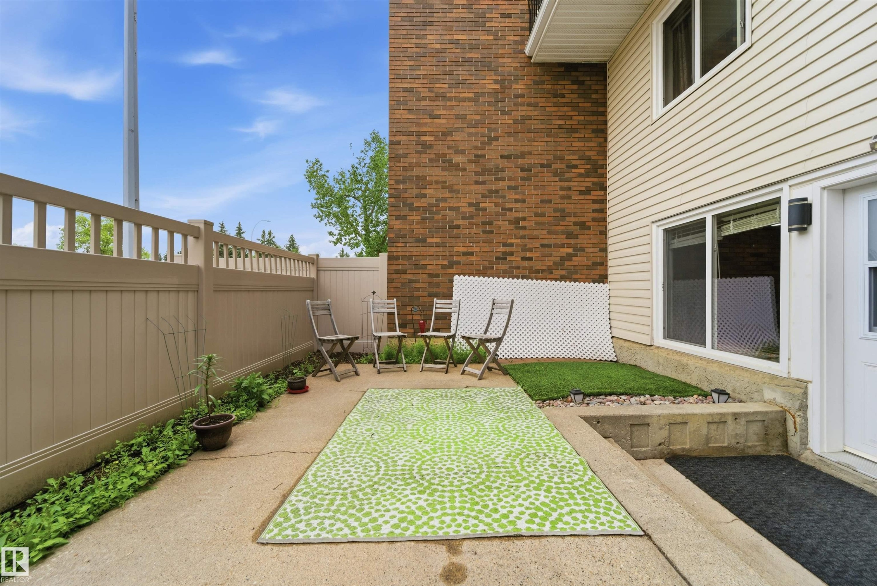 Private concrete patio area featuring a tan privacy fence and a faux grass landscaping element - #110 7805 159 St Nw, Edmonton, AB