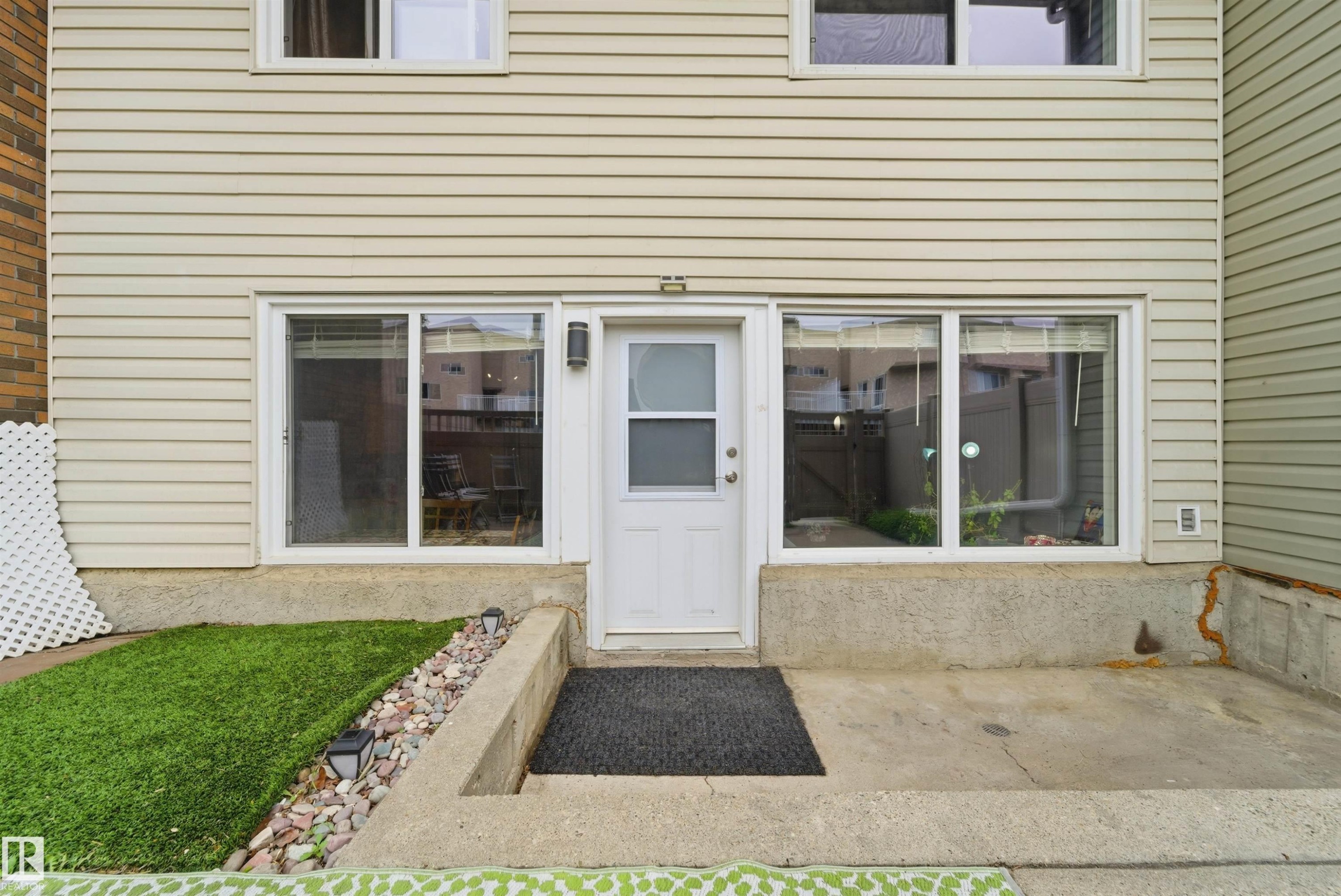 Rear entrance with white paneled door, flanked by large windows, featuring concrete patio space, and a small green lawn area with decorative rock landscaping - #110 7805 159 St Nw, Edmonton, AB