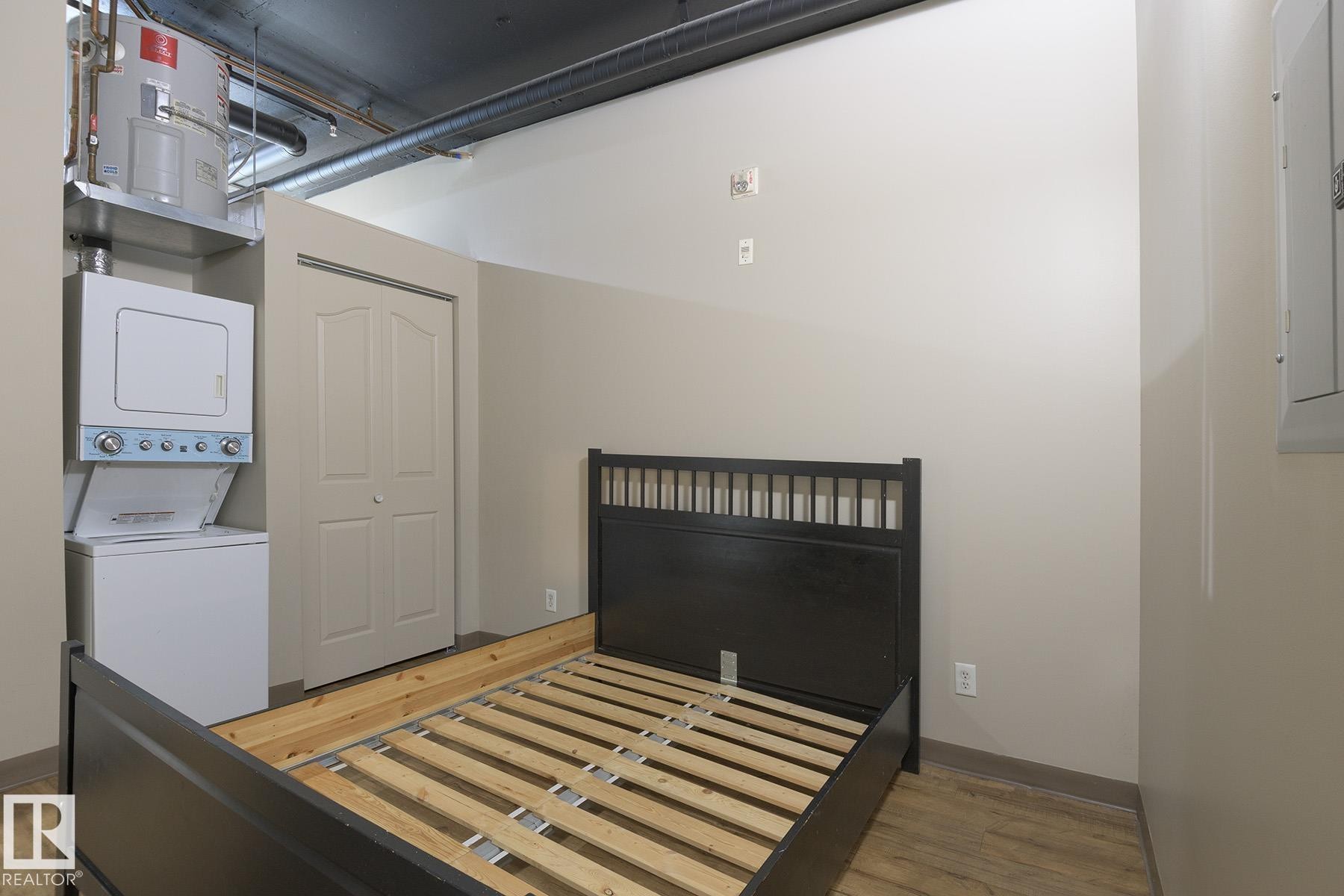 Compact laundry stack, bi-fold closet doors, exposed industrial-style ceiling ductwork, beige wall paint, and wood-finish flooring - #412 10024 Jasper Av Nw, Edmonton, AB - Indoor