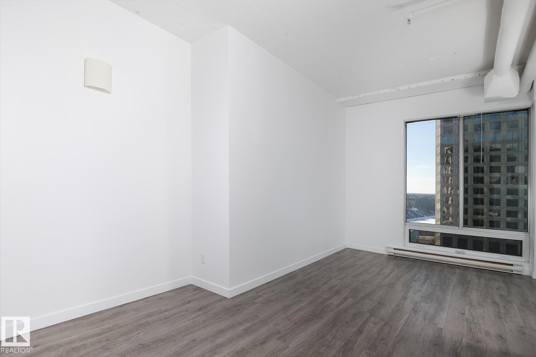 Expansive window with urban scenery, featuring wood-finish flooring, white baseboards, and a wall-mounted heating unit - 910 10024 Jasper Avenue, Edmonton, AB