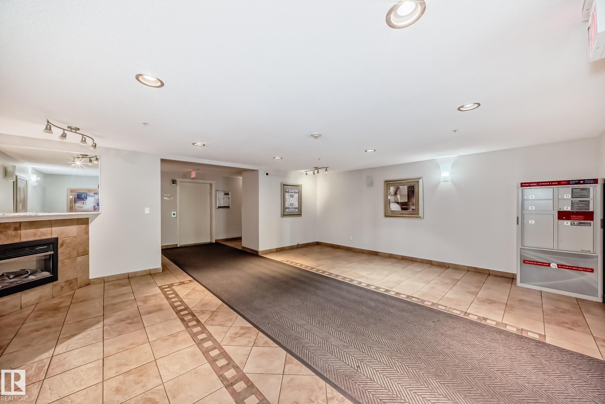 Lobby area featuring ceramic tile flooring with decorative inlay, a built-in electric fireplace with stone surround, recessed ceiling lighting, and wall-mounted light fixtures - 214 9938 104 Street, Edmonton, AB