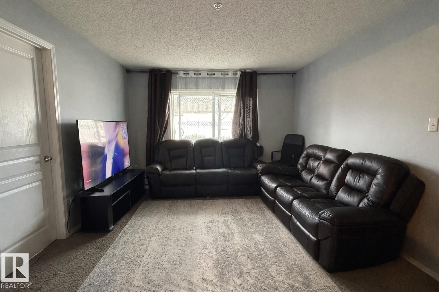 Living area featuring light gray walls, textured ceiling, a white paneled door, and a window with blinds and drapes - 133 301 Clareview Station Drive, Edmonton, AB