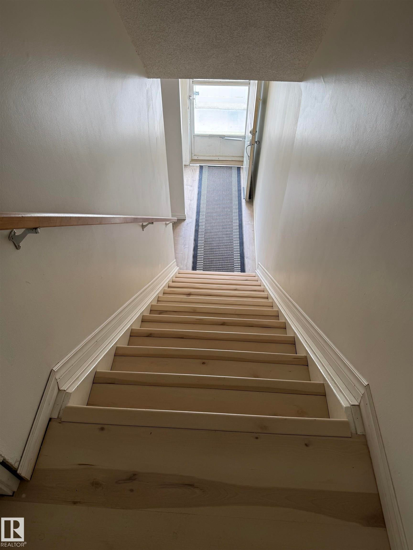 Unfinished wood staircase with a wood handrail, white wall finishes, and white baseboards, leading to a door with a window - 14537 121 Street, Edmonton, AB