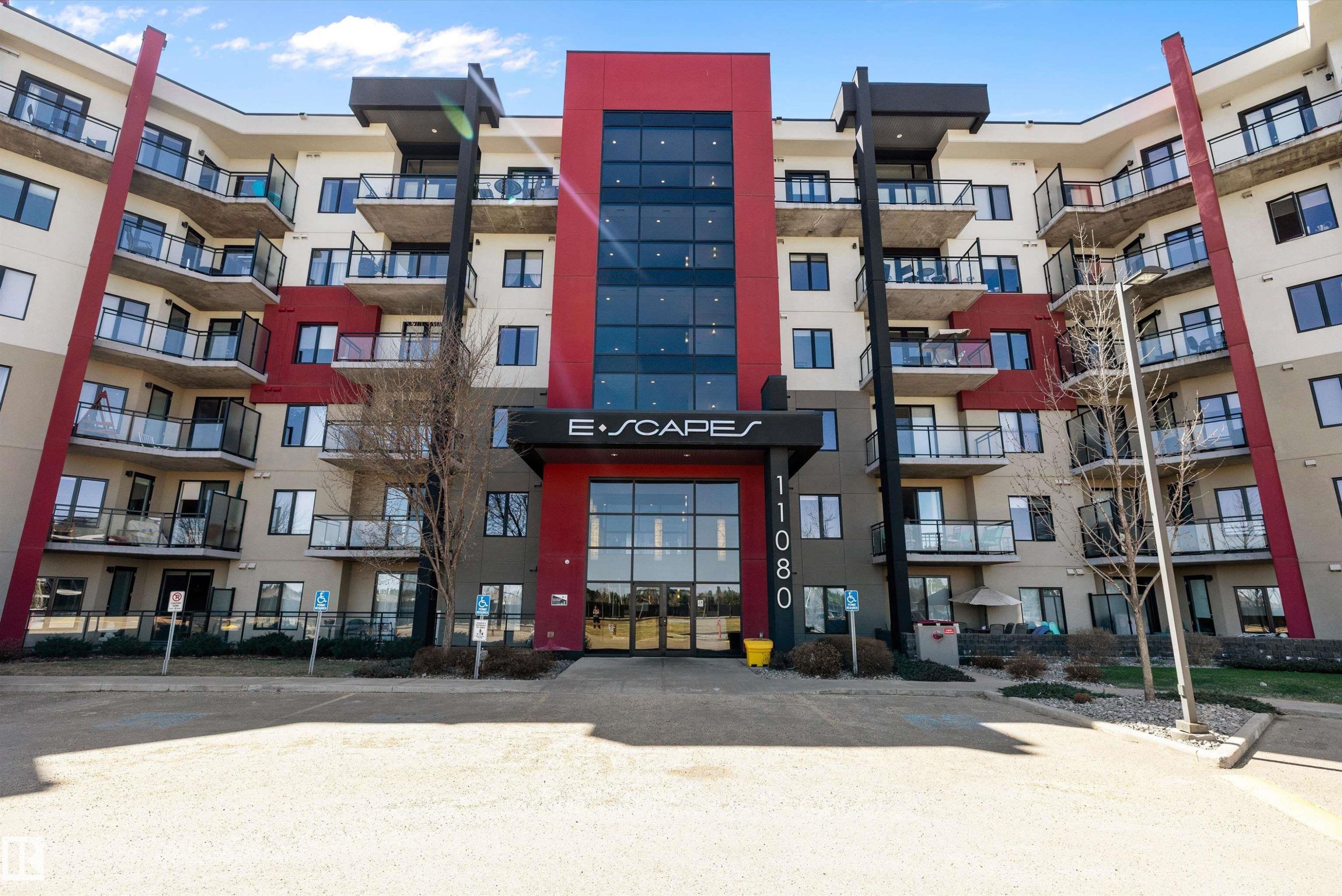 Modern residential building with a prominent central entrance, multiple levels of private balconies featuring glass railings, red and neutral-toned facade accents, and a landscaped perimeter. - 307 11080 Ellerslie Rd Sw, Edmonton, AB